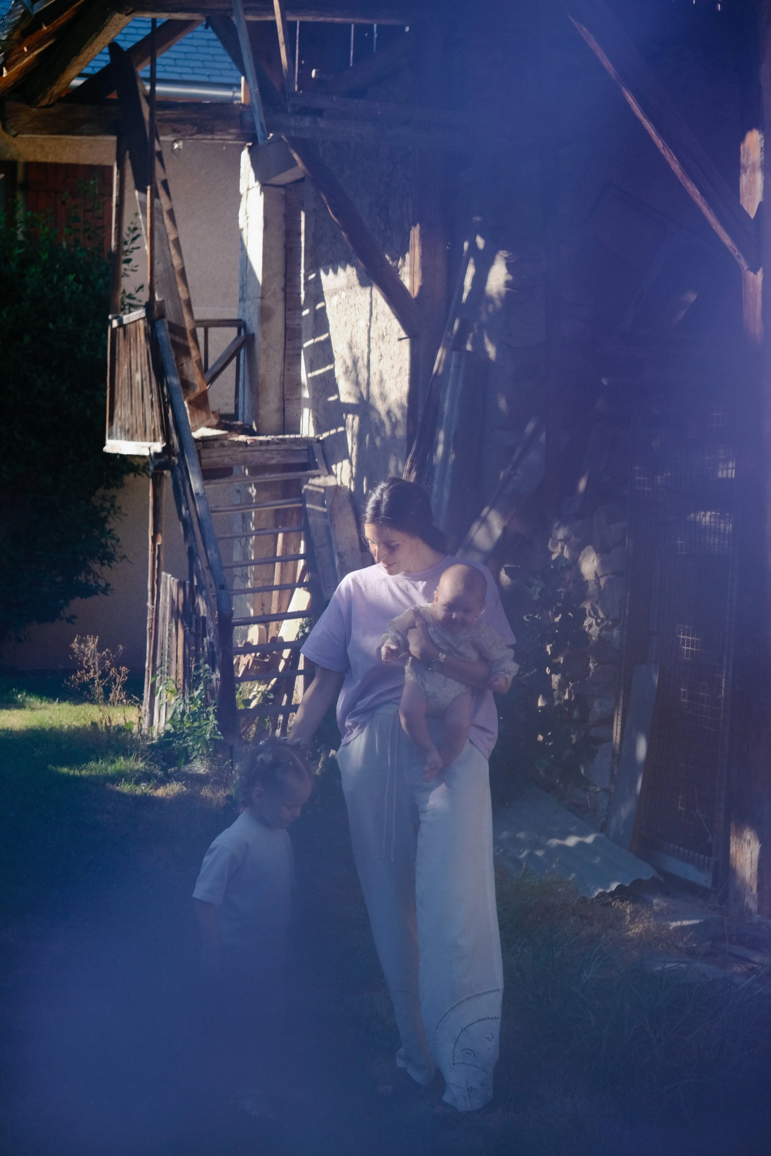 Une femme tenant un bébé et deux enfants en vêtements blancs dans un jardin avec une structure en bois en ruine et un arbre vert.