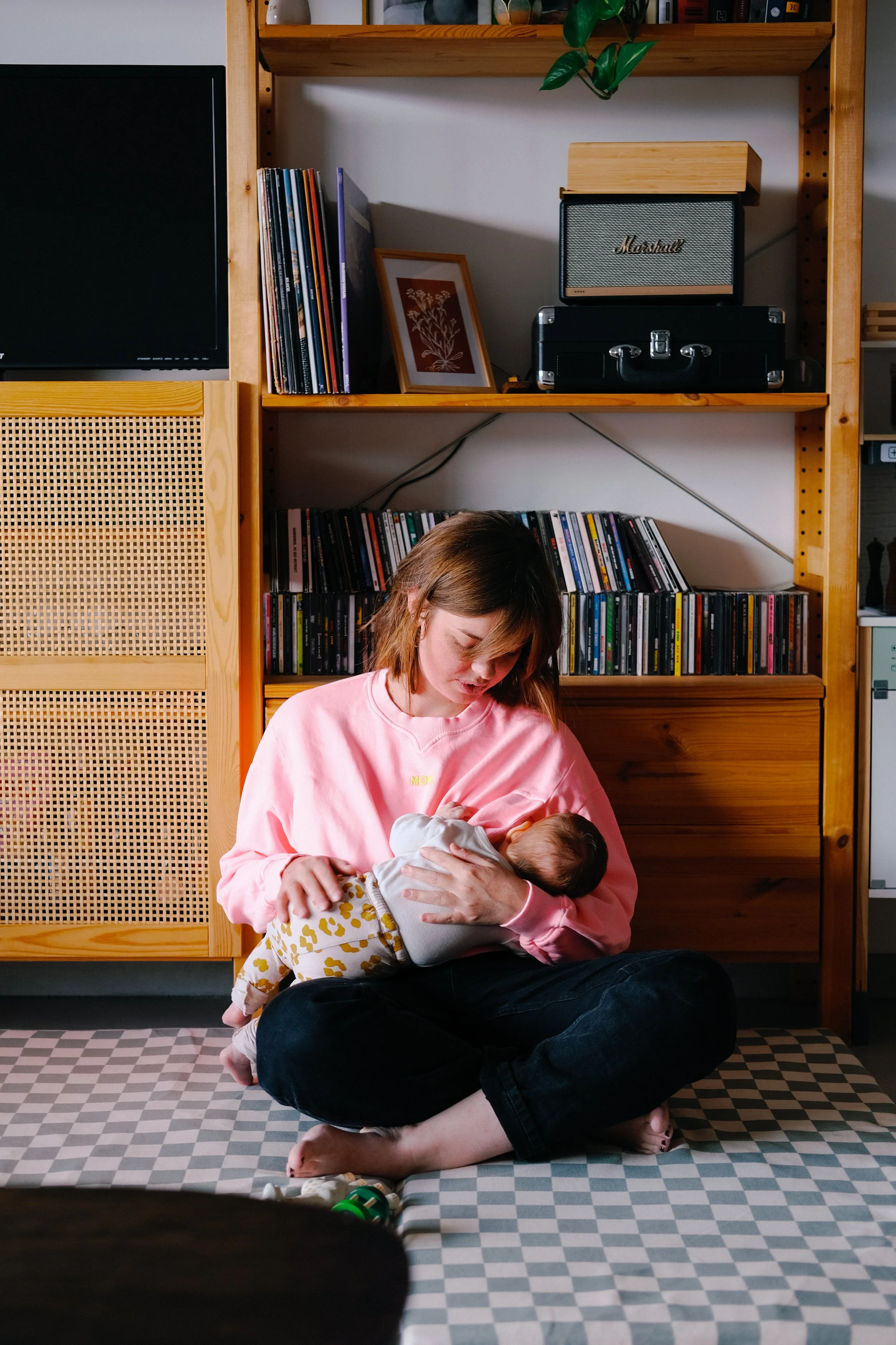 Une jeune femme en sweat rose tient un bébé dans ses bras. Ils sont assis par terre devant une étagère en bois contenant des livres, un cadre photo, une petite enceinte Marshall, et d'autres objets. La femme regarde doucement le bébé qui semble dormi