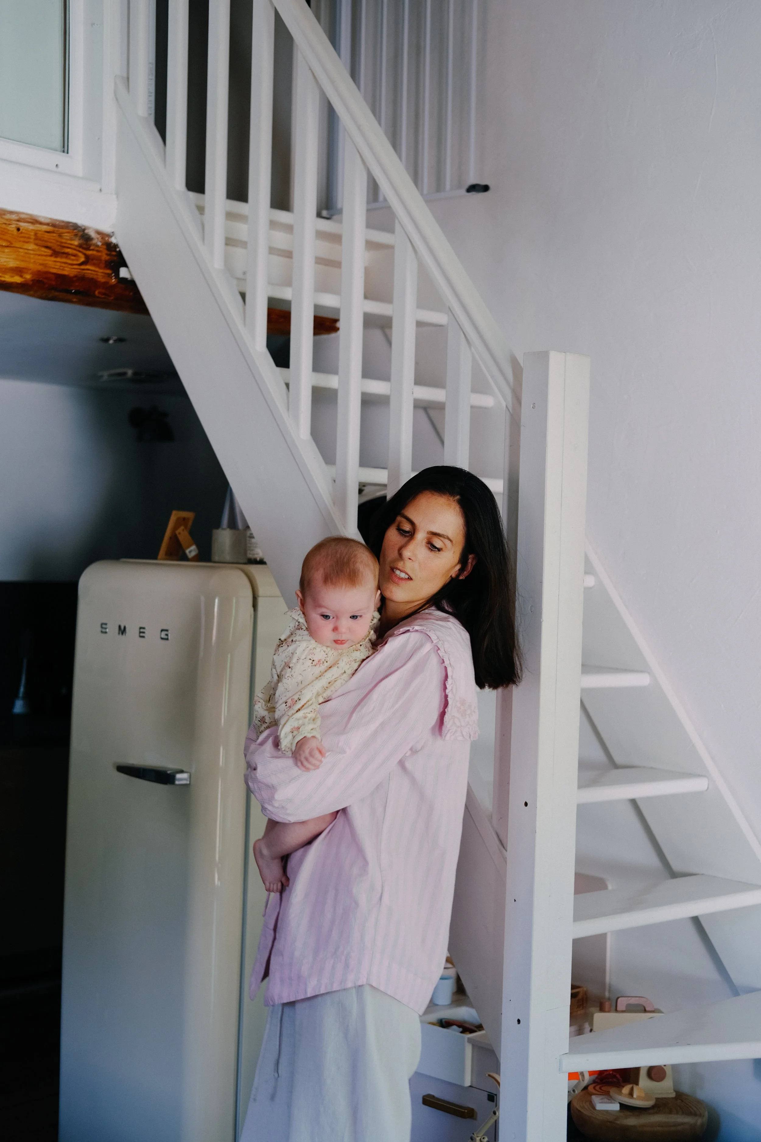 Une femme tenant un bébé dans ses bras dans une maison, avec un escalier blanc en arrière-plan.