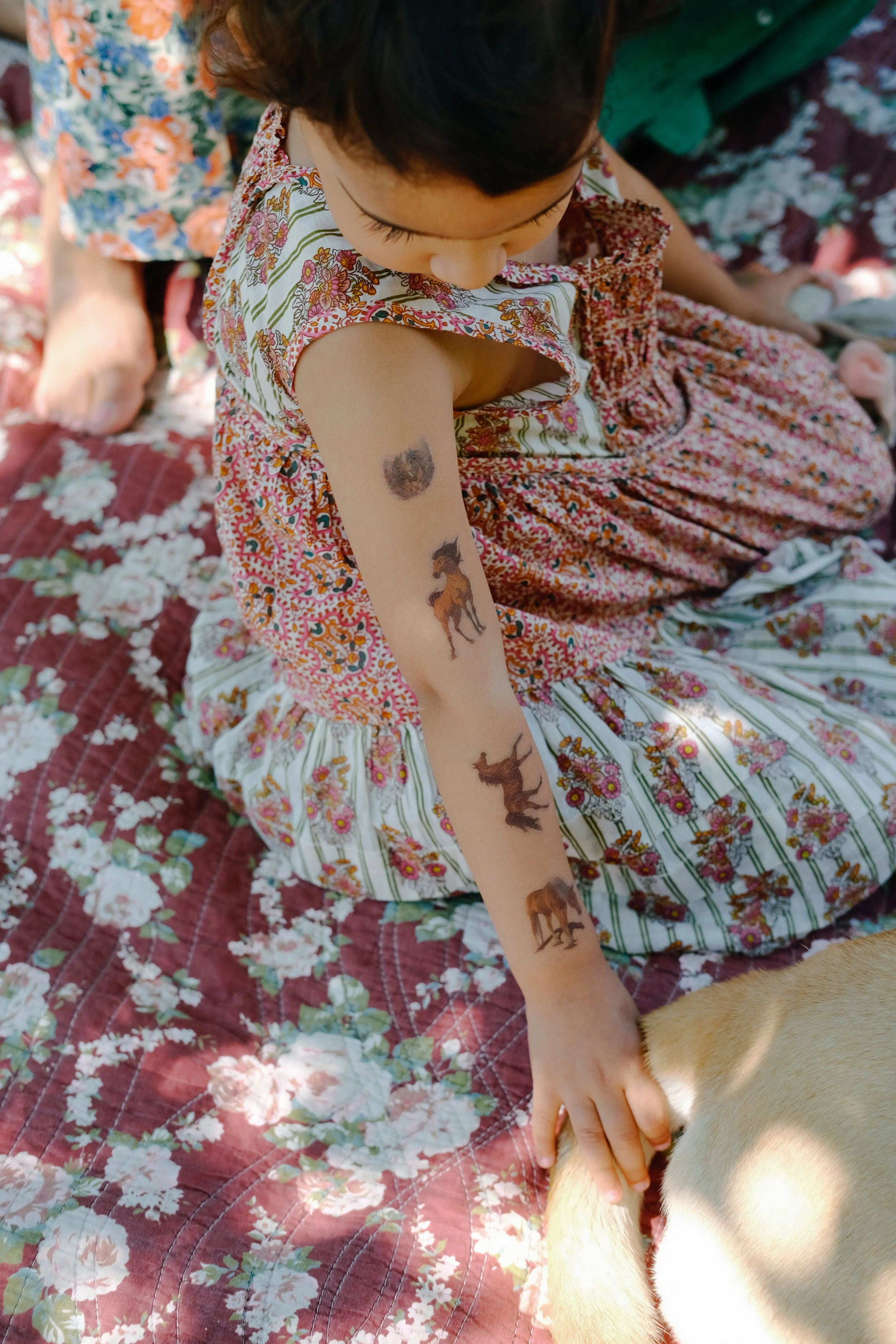 Une jeune fille avec des tatouages de chevaux sur le bras, assise sur une couverture à fleurs, caressant un chien de couleur claire.