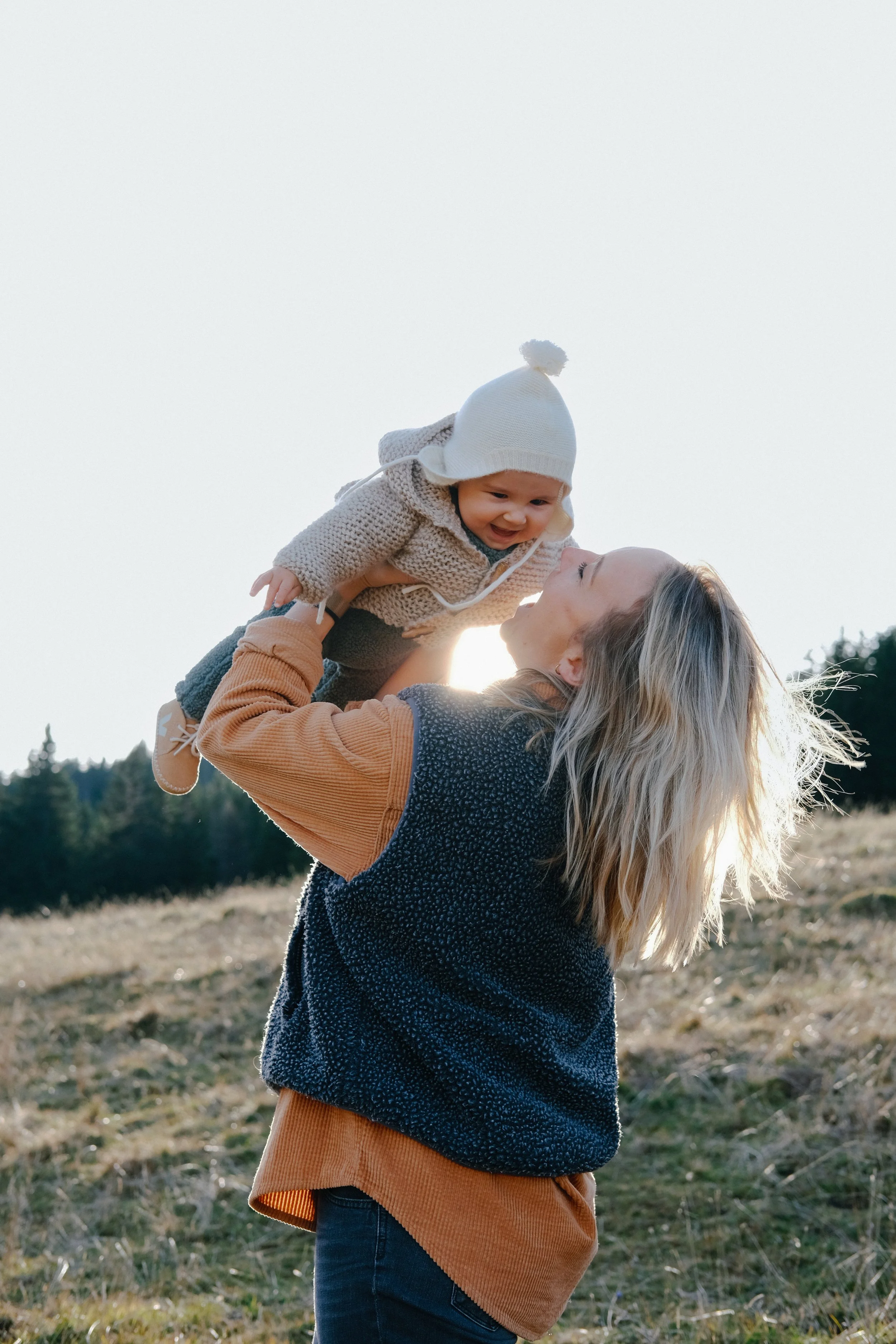 Une femme blonde en pull noir et veste orange tenant un bébé dans ses bras dans un champ en plein air.