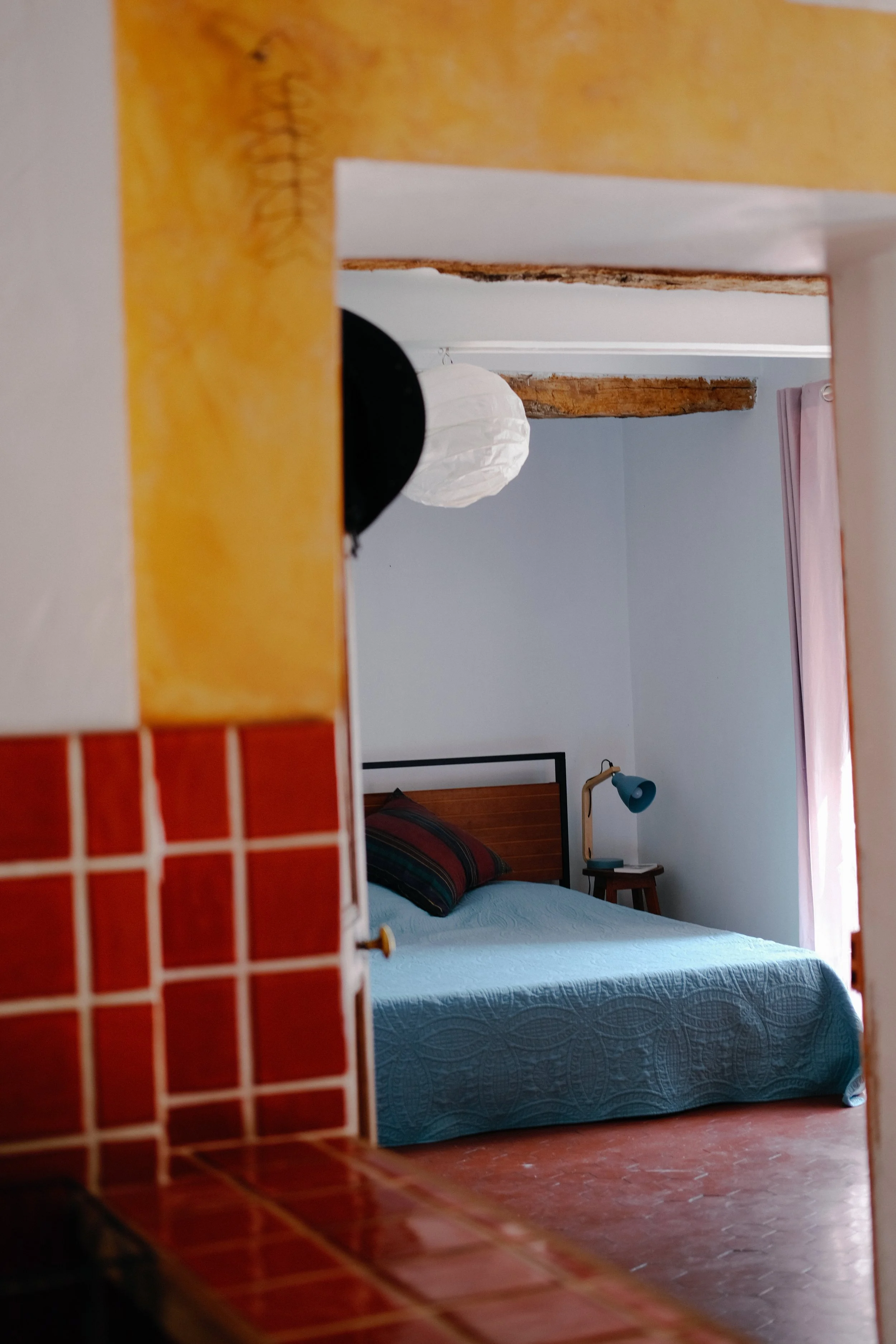 Vue de la chambre d'une maison, avec un lit en bois, une lampe de chevet, une fenêtre avec rideau rose, un mur blanc, et un plafond avec poutres en bois
