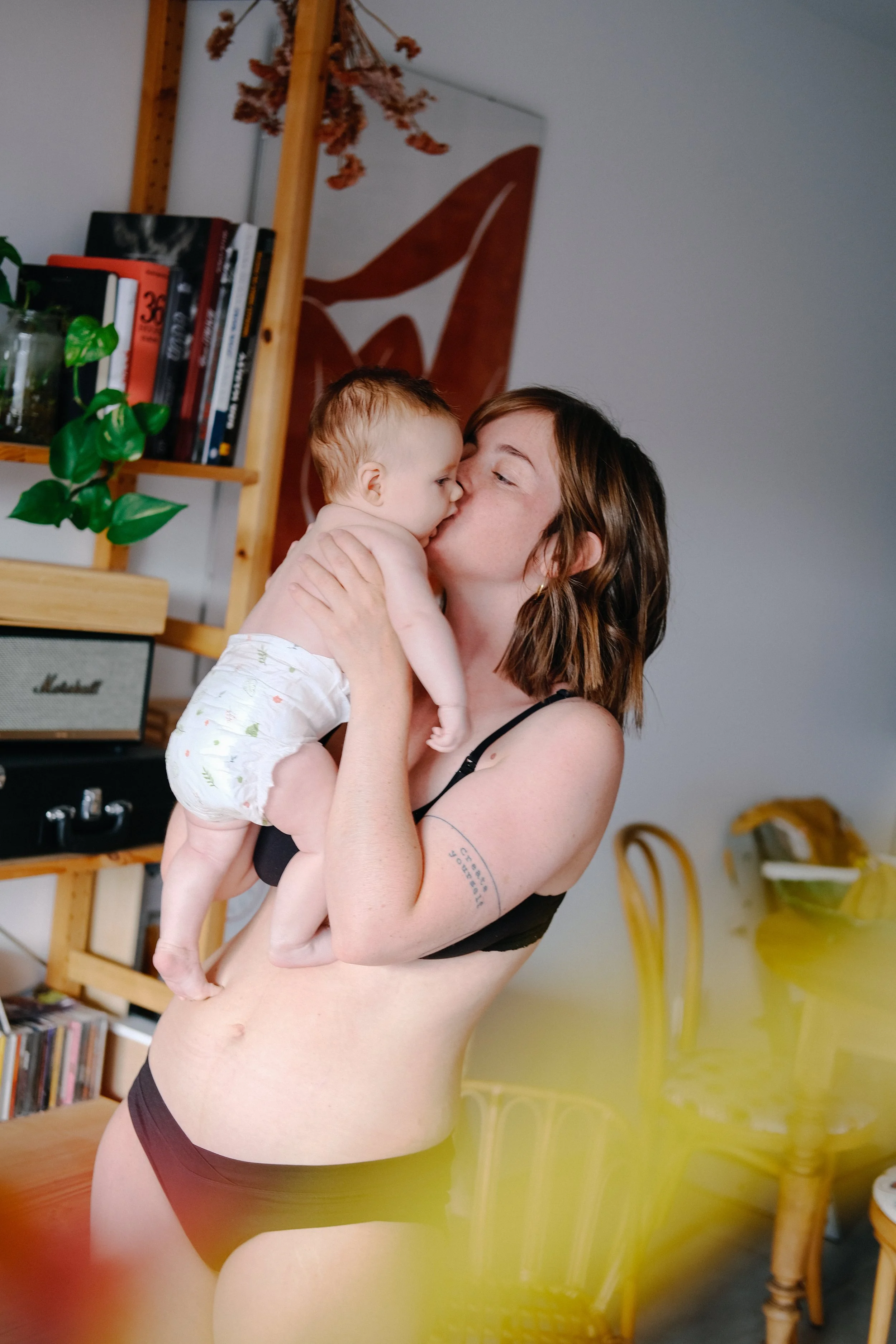 Une femme debout, tenant un bébé dans ses bras, dans une pièce avec un mur gris, une étagère en bois avec des livres et des plantes, et un tableau abstrait. La femme porte un soutien-gorge noir et des sous-vêtements marrons.