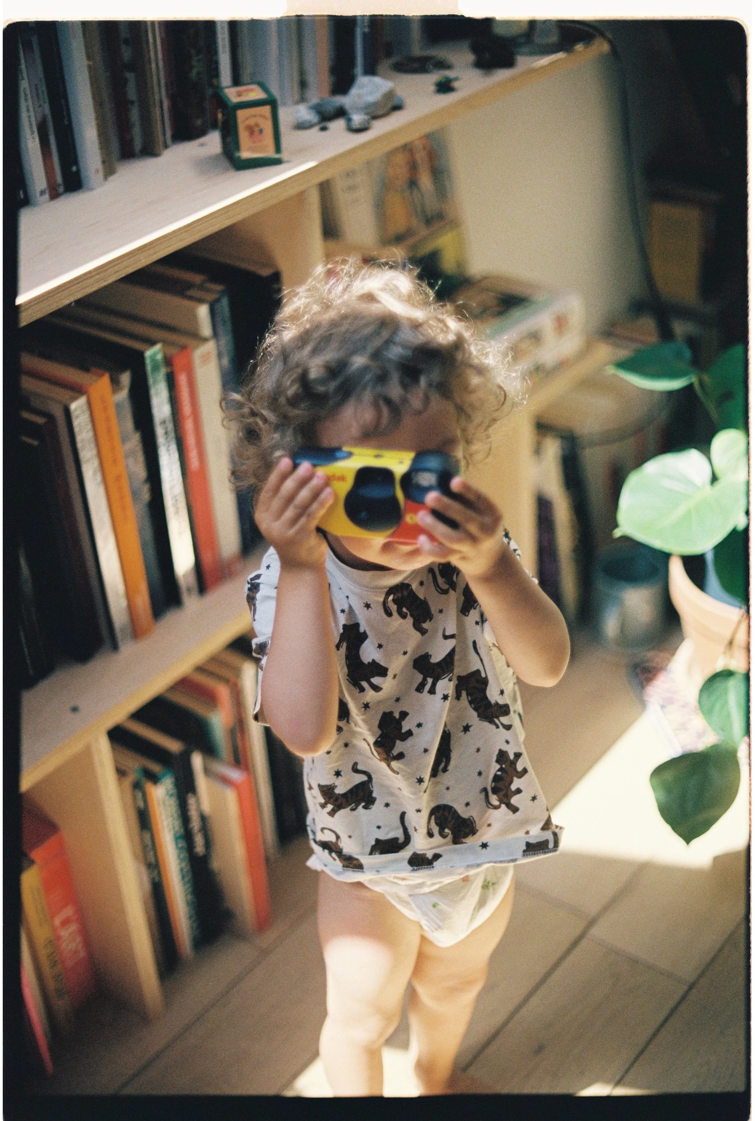 Un enfant debout dans une bibliothèque, portant un t-shirt blanc avec des léopards, tenant un appareil photo et couvrant ses yeux avec.