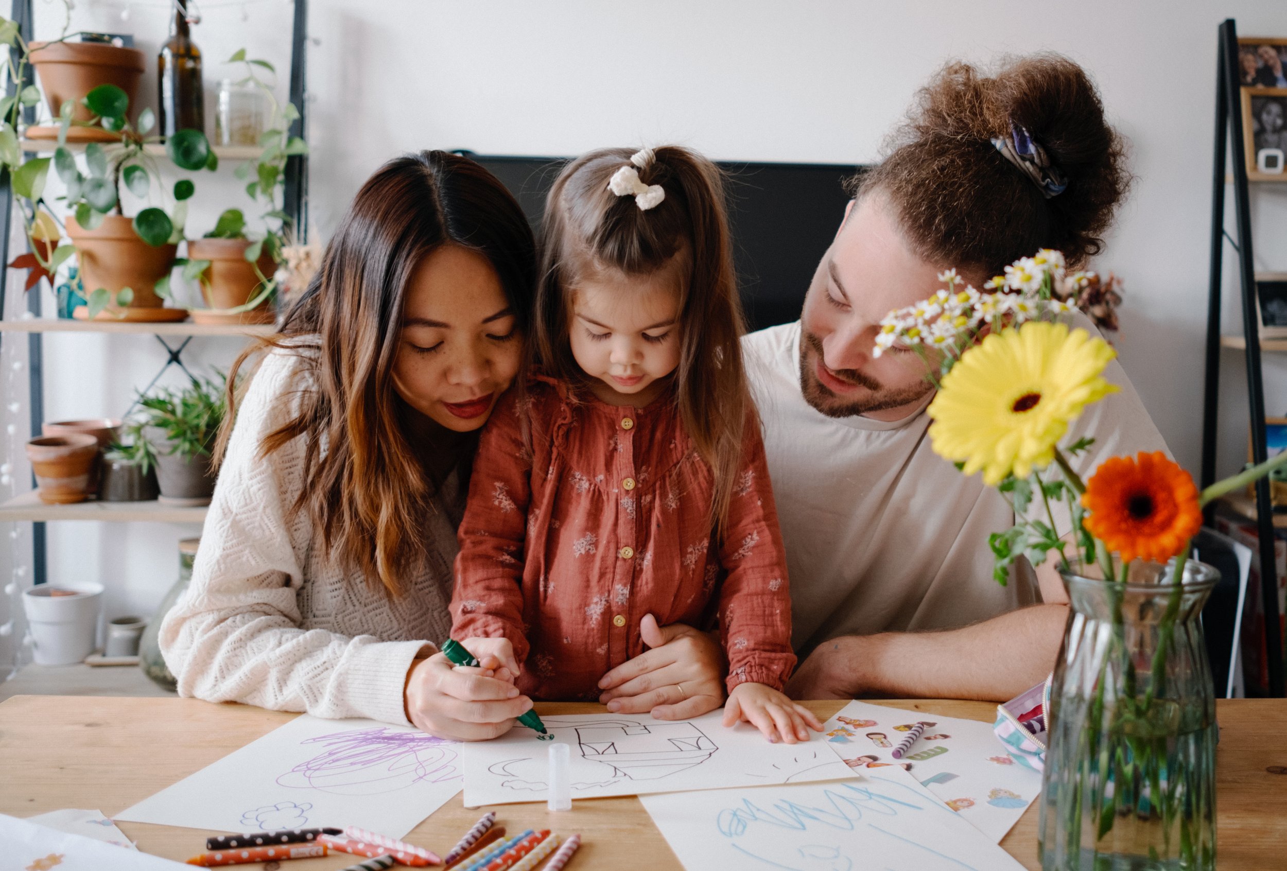 Une famille compose d'une mère, d'un père et d'une fille sont assis autour d'une table de bois, en train de dessiner et d'écrire sur des feuilles. La mère aide la petite fille à colorier. La table est également couverte de crayons, de feutres et de d