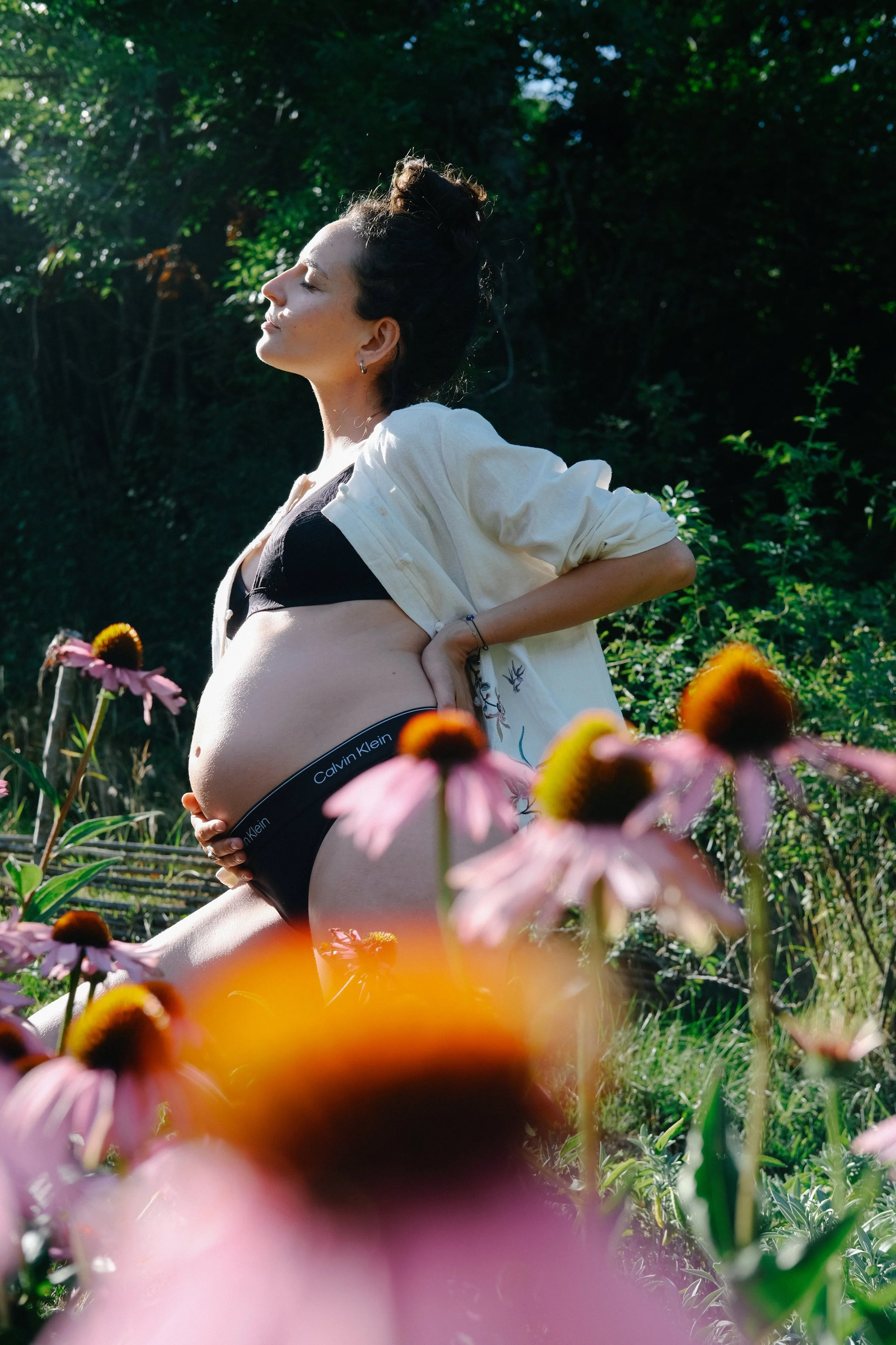 Une femme nue tenant son propre ventre arrondi, portant un débardeur noir et une veste beige, dans un jardin fleuri avec des fleurs roses au premier plan, en pleine lumière naturelle.