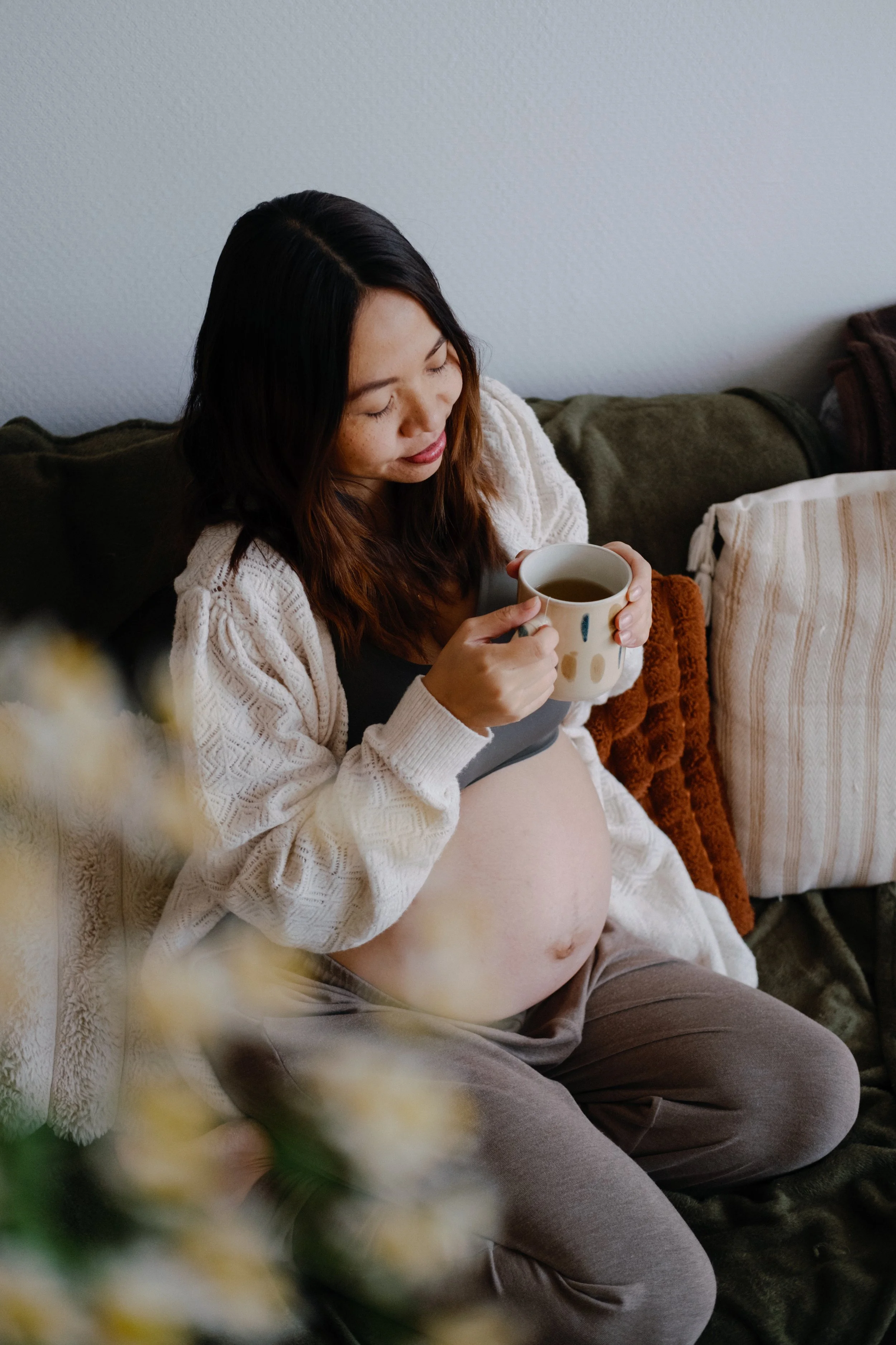 Femme enceinte assise sur un canapé, tenant une tasse, avec des coussins et des vêtements confortables autour.