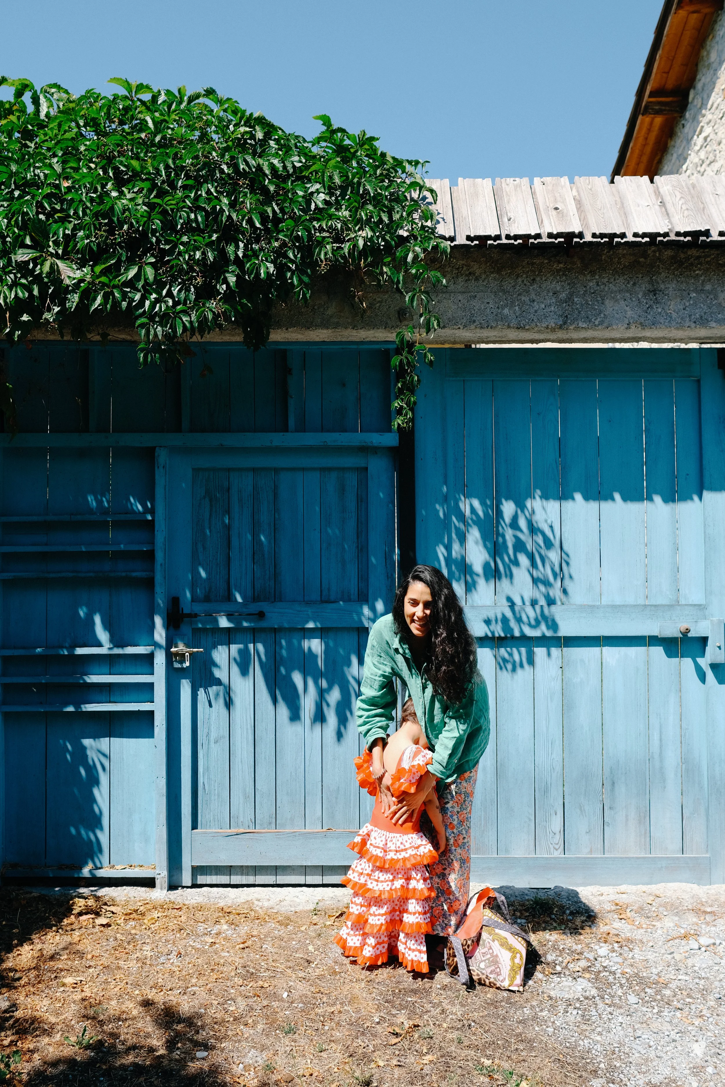 Une femme avec de longs cheveux noirs, portant une veste verte, et une jeune fille en robe à volants orange, se tenant devant une clôture bleue en bois, avec une maison et un ciel bleu en arrière-plan.