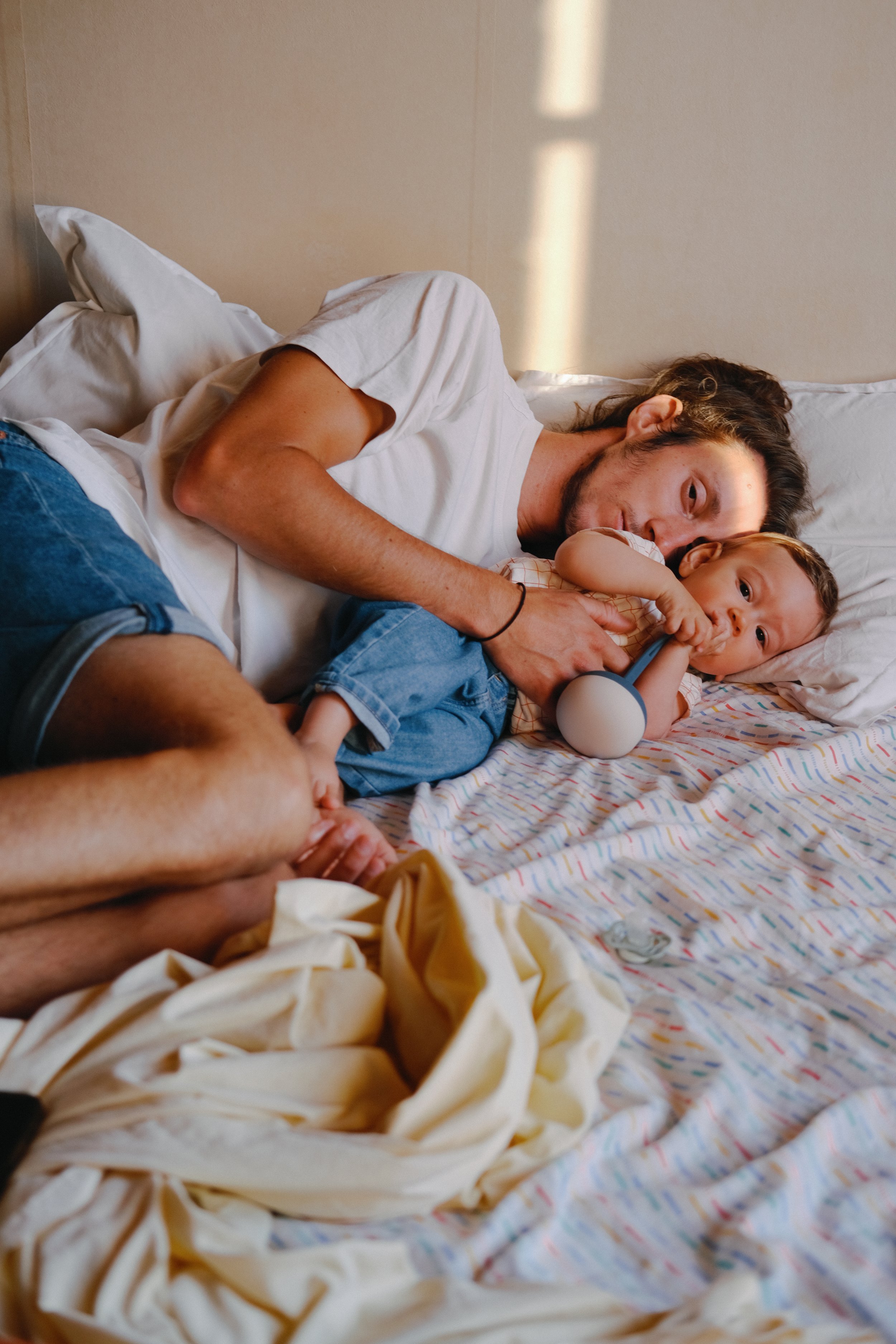 Un homme et un jeune garçon allongés tous deux sur un lit, en train de se détendre ou de faire une sieste, avec des oreillers blancs et une couverture à rayures colorées.