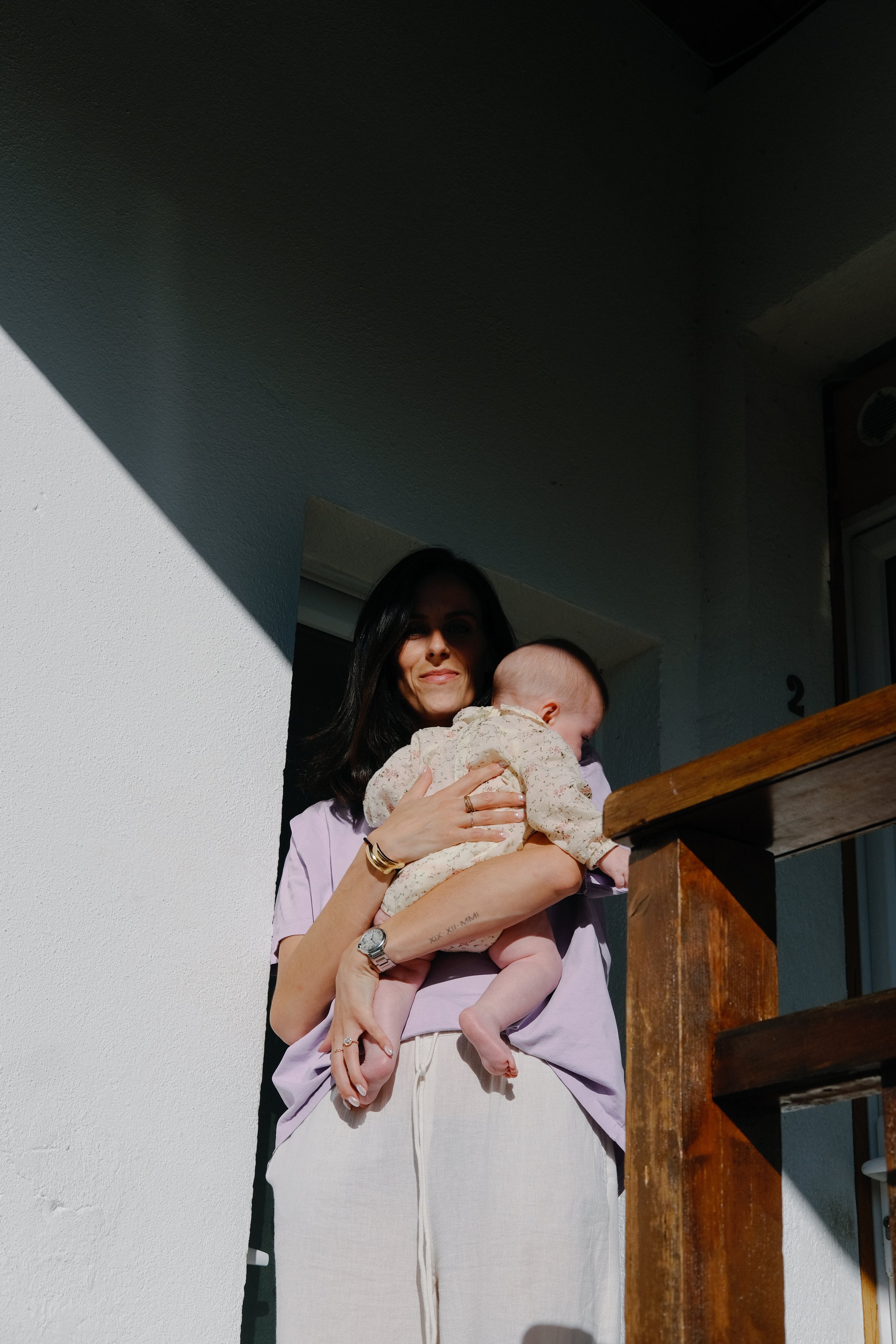 Une femme tenant un bébé sur une terrasse ensoleillée avec un mur blanc et un balcon en bois.