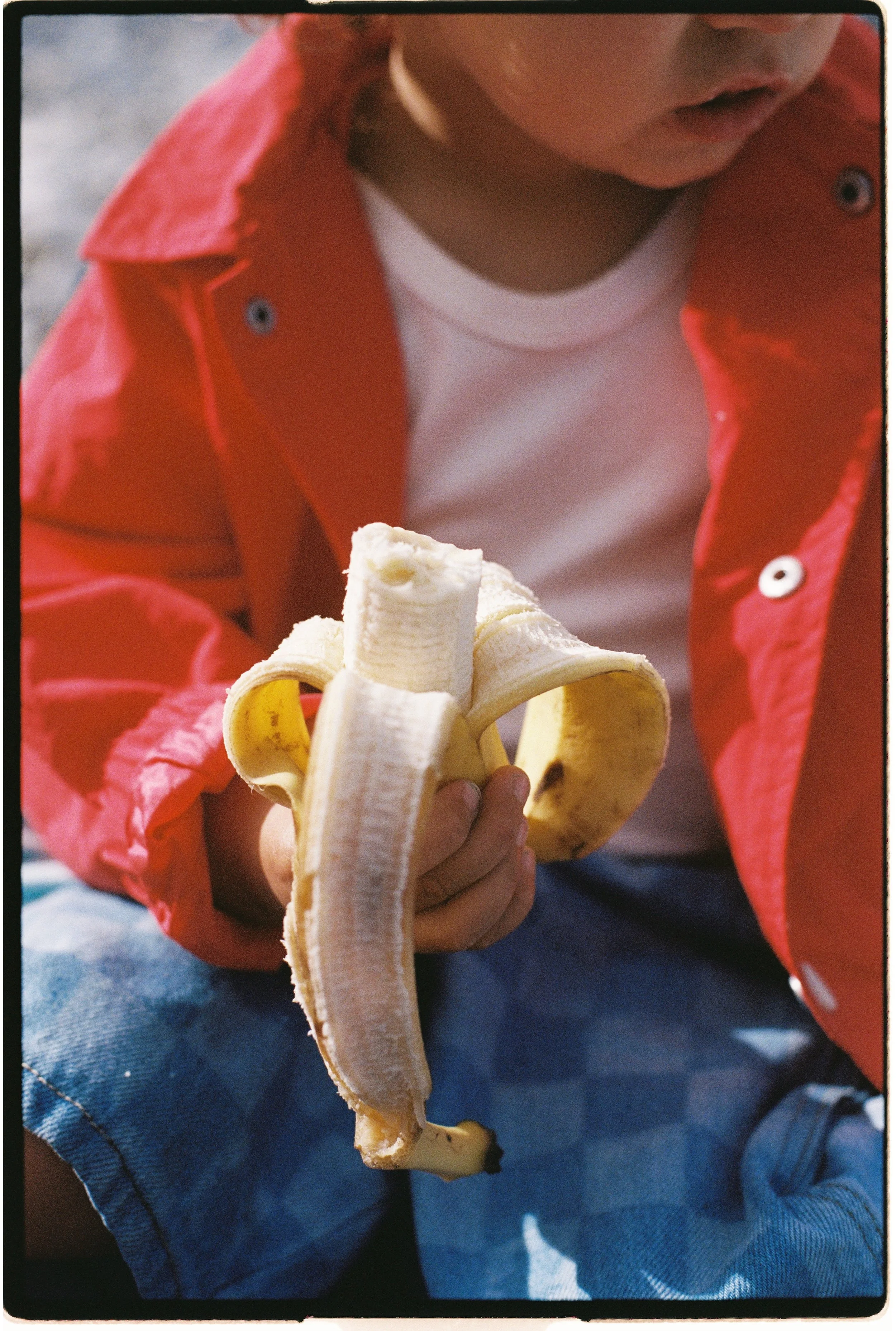 Un enfant portant une veste rouge et un t-shirt blanc tient une banane pelée.