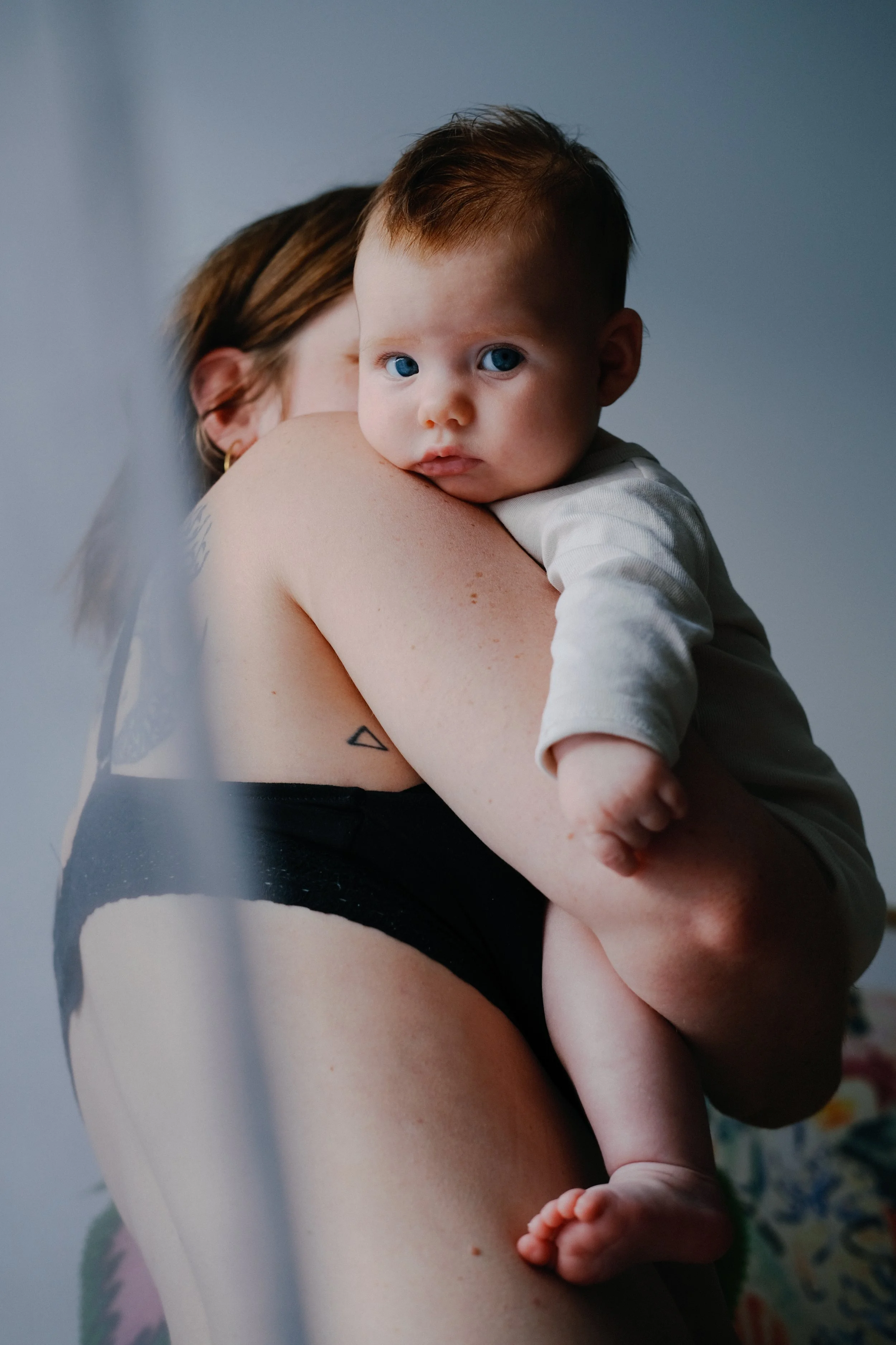 Une jeune mère tient un enfant dans ses bras, le regardant fixement. L'enfant demeure appuyé sur son épaule, semblant écouter ou regarder quelque chose. La scène est intime et douce, avec un arrière-plan neutre et quelques motifs colorés à la base.
