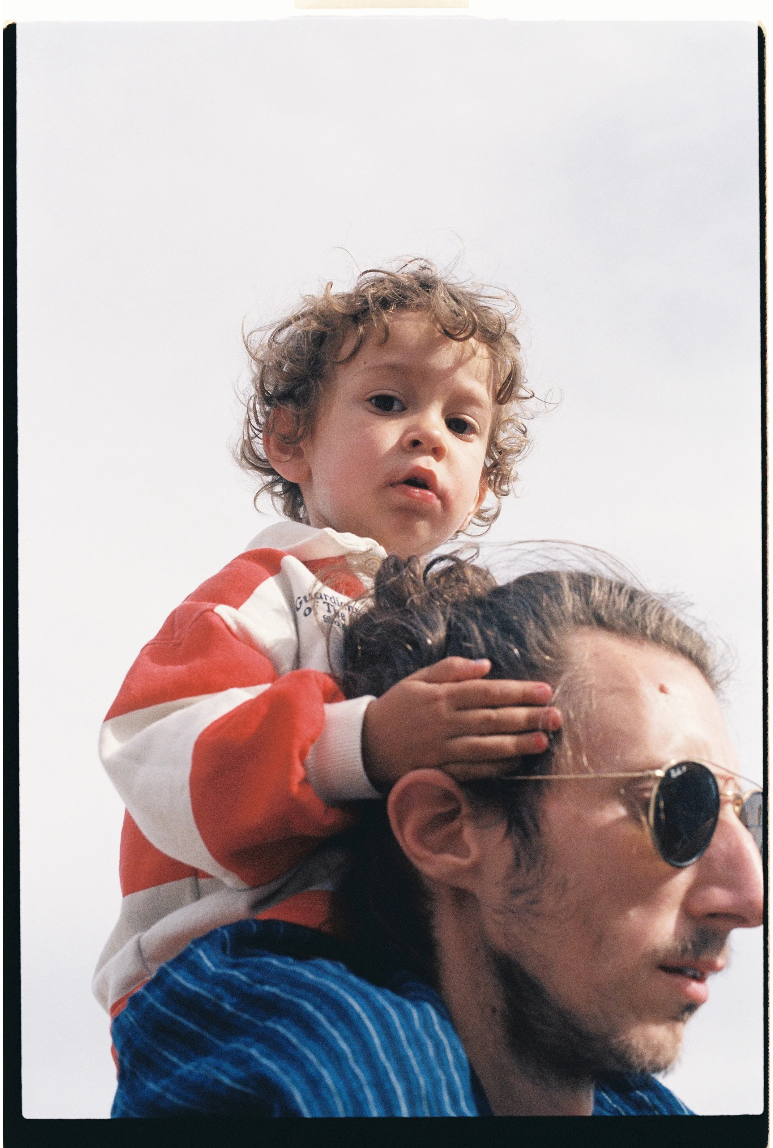 Un garçon aux cheveux bouclés, assis sur les épaules d'un adulte, portant un sweat rouge et blanc, regarde vers le haut avec un visage curieux ou pensif. L'adulte porte des lunettes de soleil et une chemise rayée. Le fond est clair et sans détails sp
