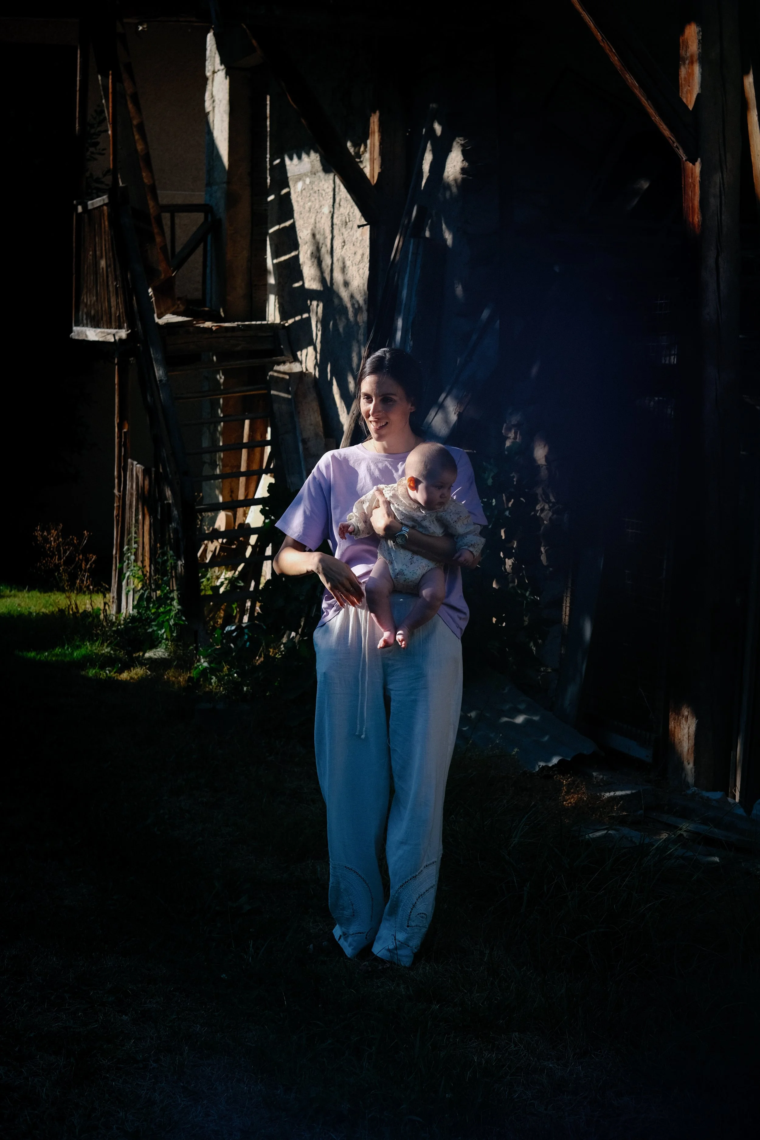 Une femme tenant un bébé devant une vieille structure en bois avec des ombres et des rayons de lumière.