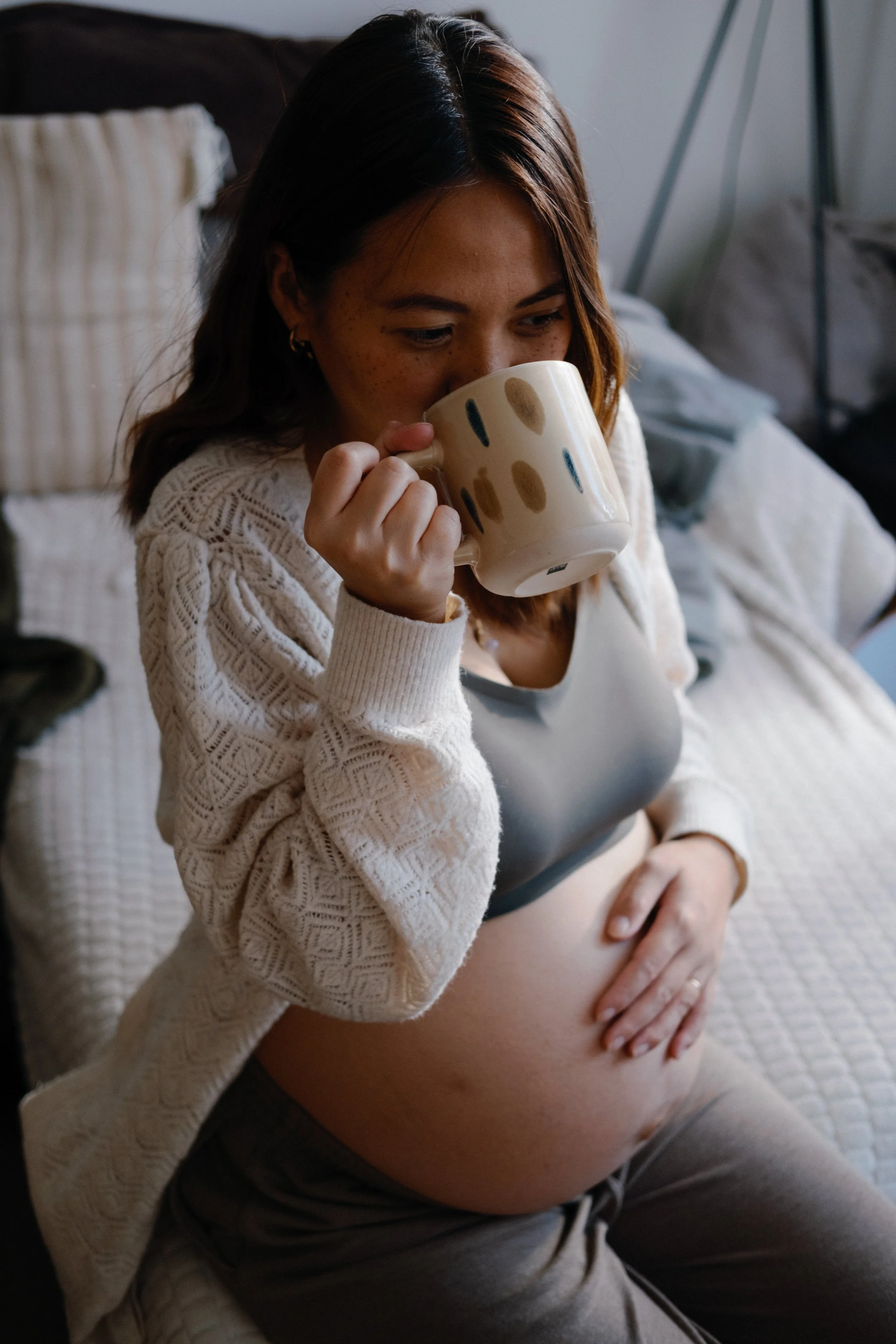 Femme enceinte prenant une tasse, portant un haut gris et un cardigan beige, assise sur un lit, dans une pièce lumineuse.