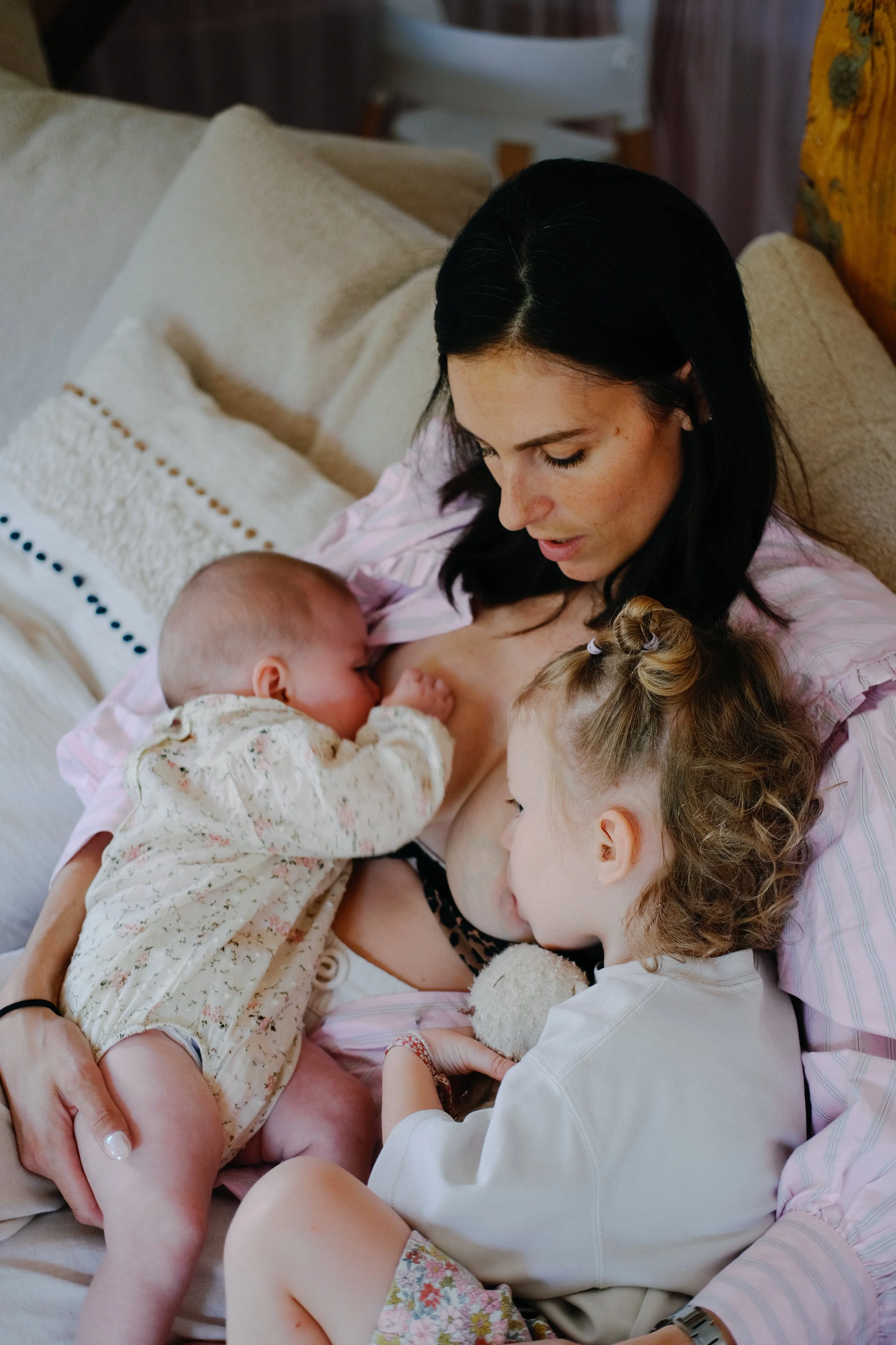 Une femme avec des cheveux noirs nourrissant deux jeunes enfants, un bébé et un tout-petit, dans une pièce chaleureuse, assise sur un canapé beige.