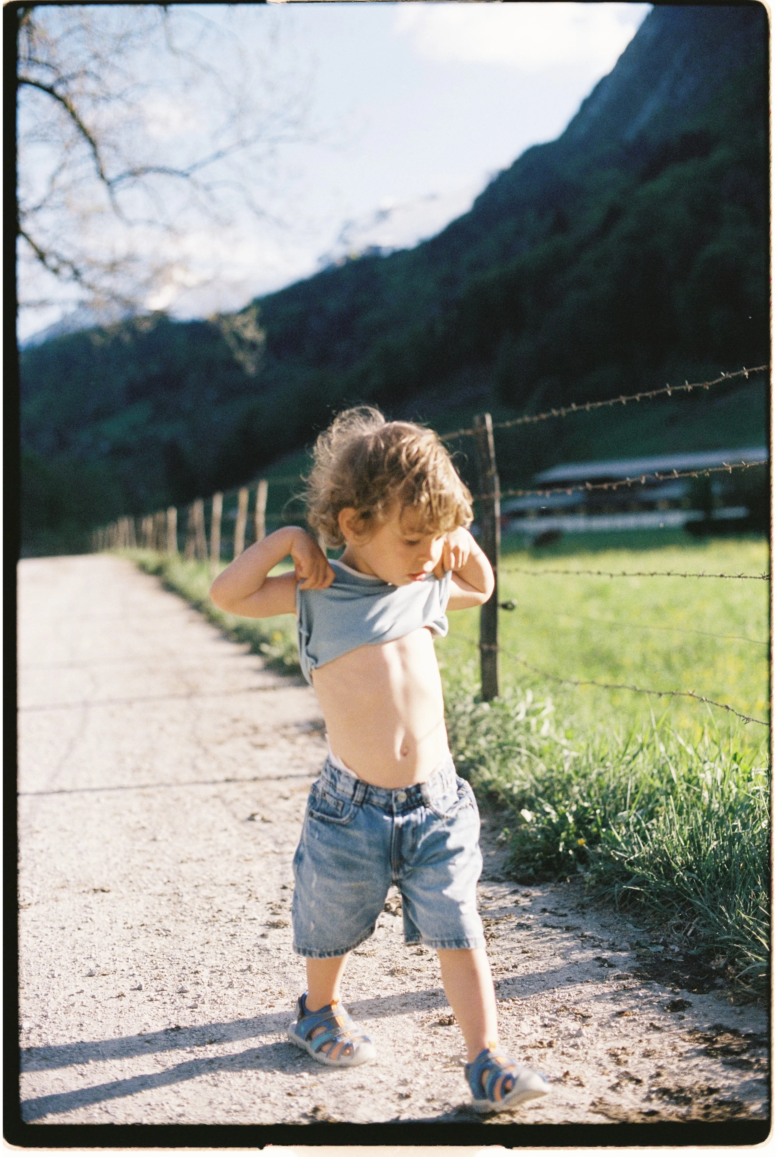Un enfant marche sur un chemin en bordure d'une prairie entourée d'une clôture, avec des montagnes en arrière-plan, en plein jour, vêtue d'un t-shirt gris, de jeans et de sandales.