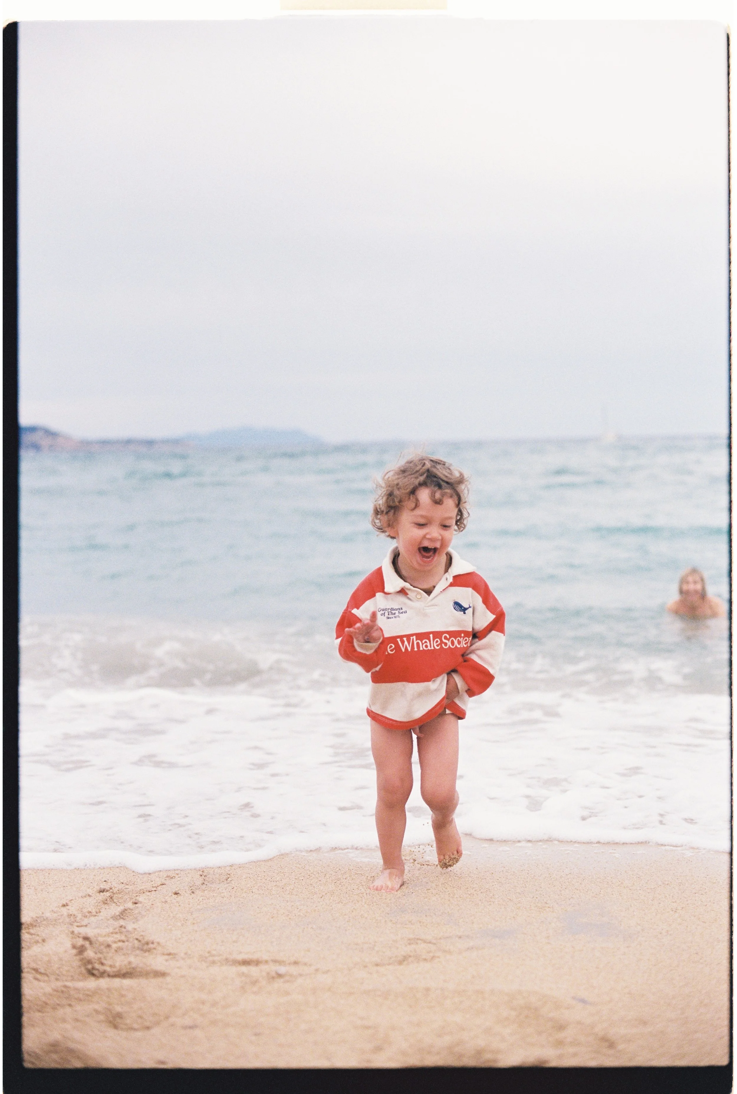 Un enfant souriant court sur la plage près de la mer, portant un sweat à rayures rouges et blanches et pas de vêtements en dessous, avec d'autres personnes à l'arrière-plan dans l'océan.