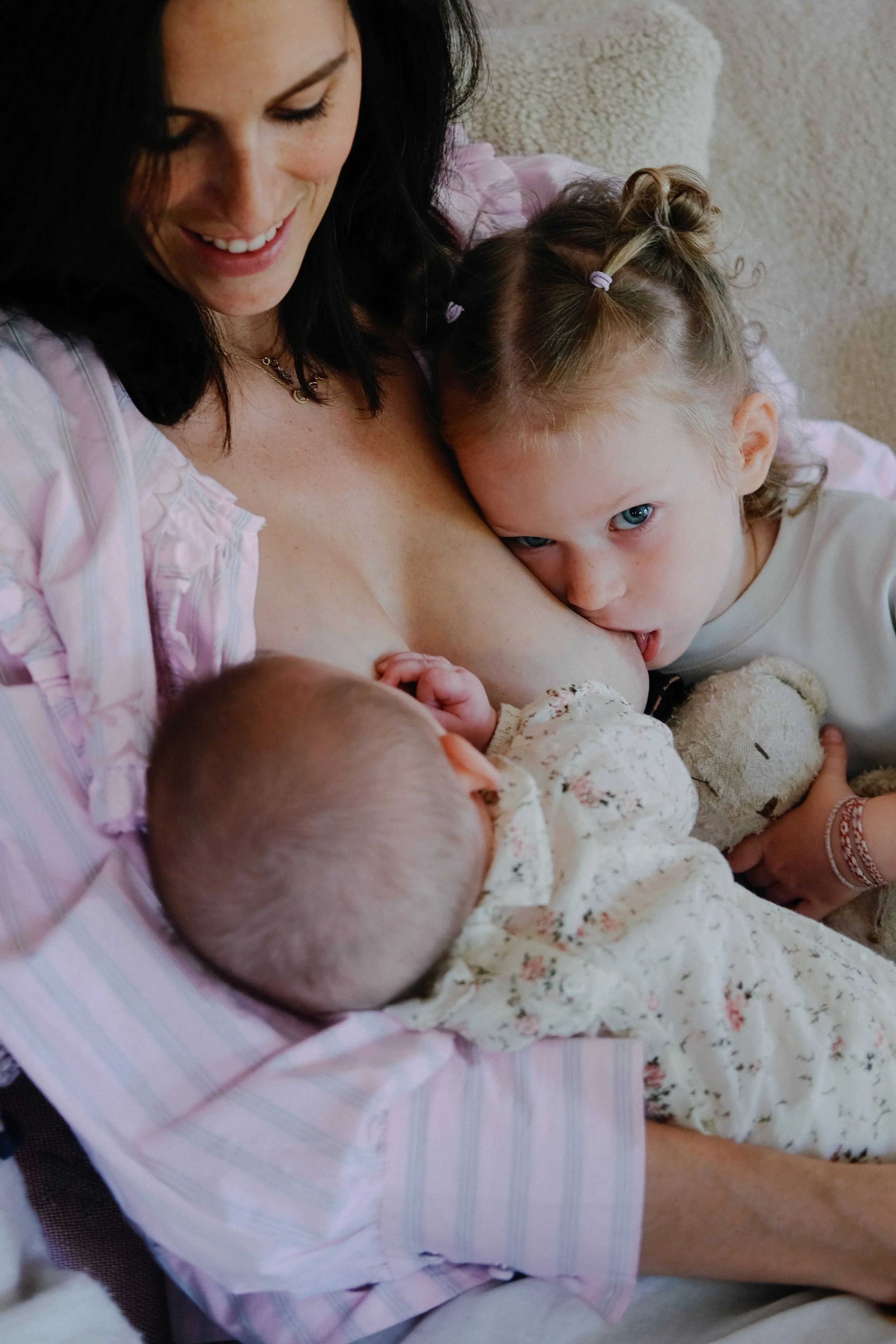 Une femme avec deux jeunes enfants, une fille à ses côtés, un bébé dans ses bras, dans une ambiance chaleureuse dans une maison.
