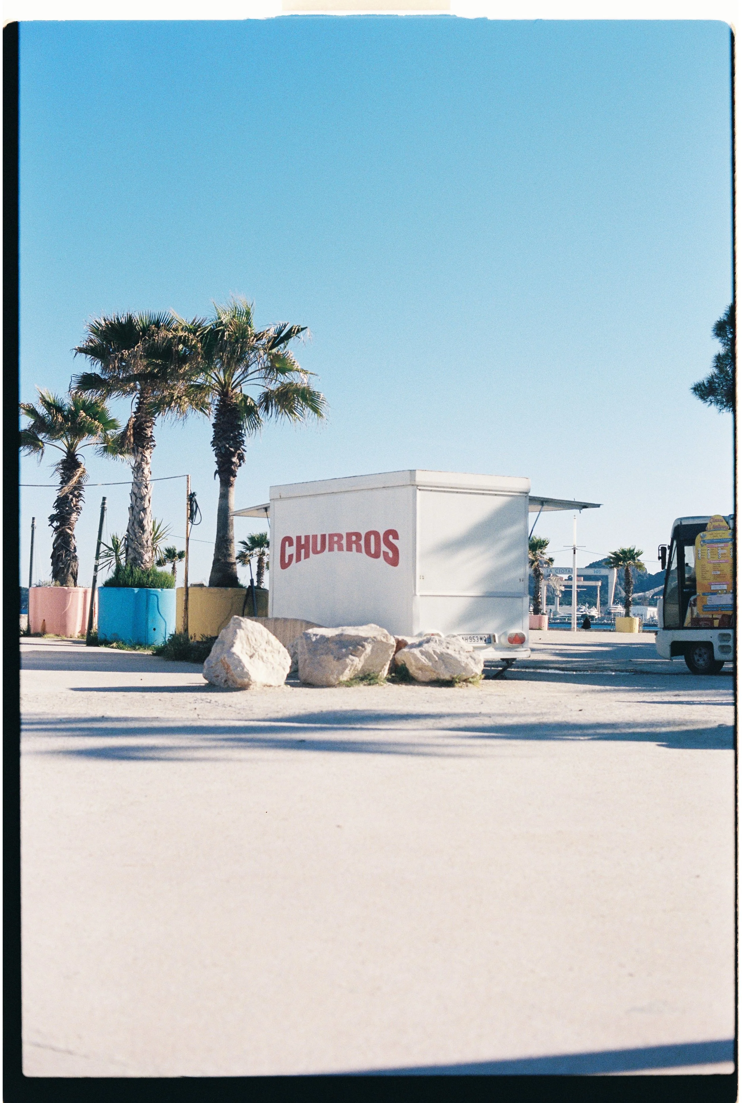 Une scène en bord de mer avec des palmiers, un stand de churros, et des rochers sur le sol.