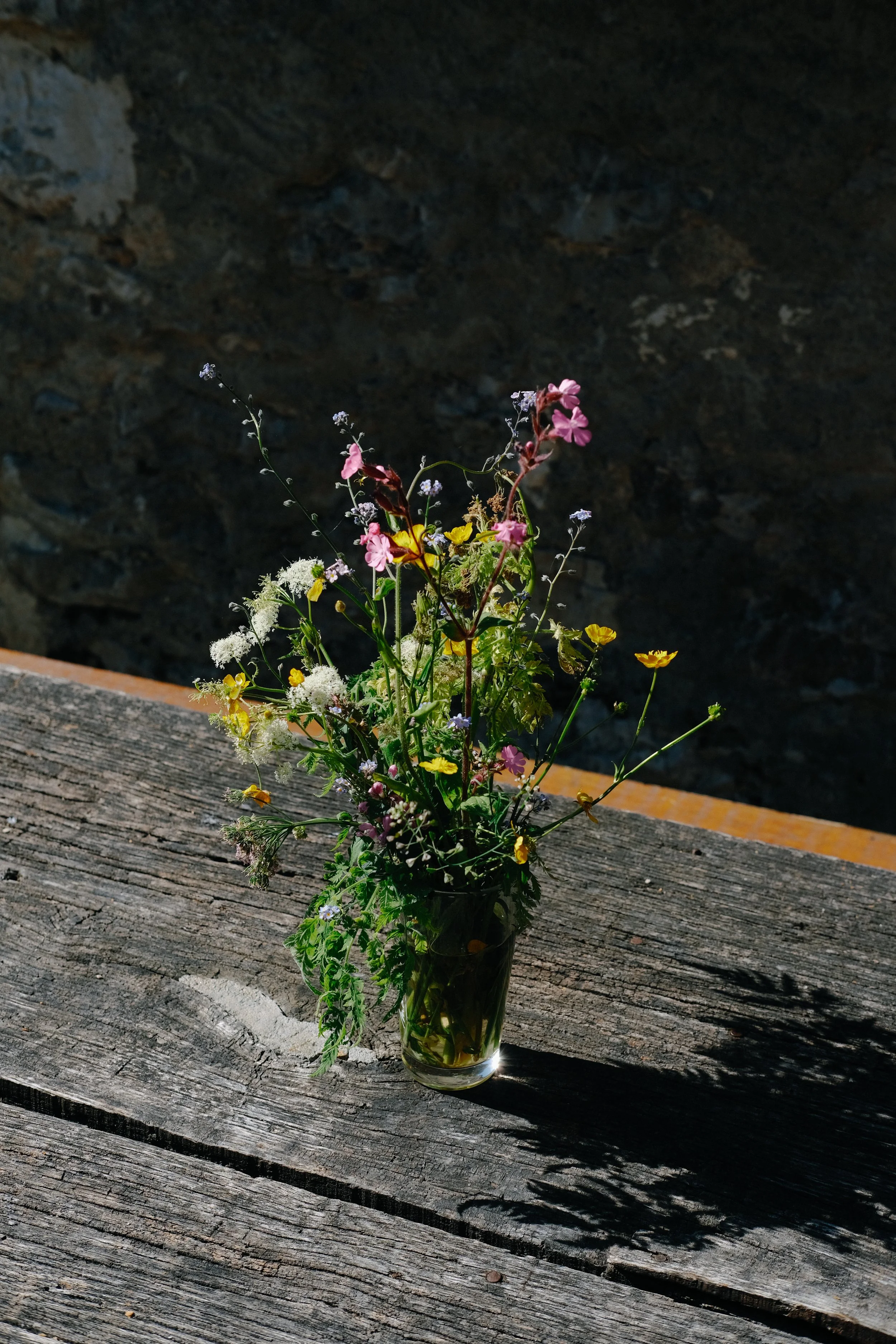 Un bouquet de fleurs sauvages dans un vase en verre sur un banc en bois en extérieur.
