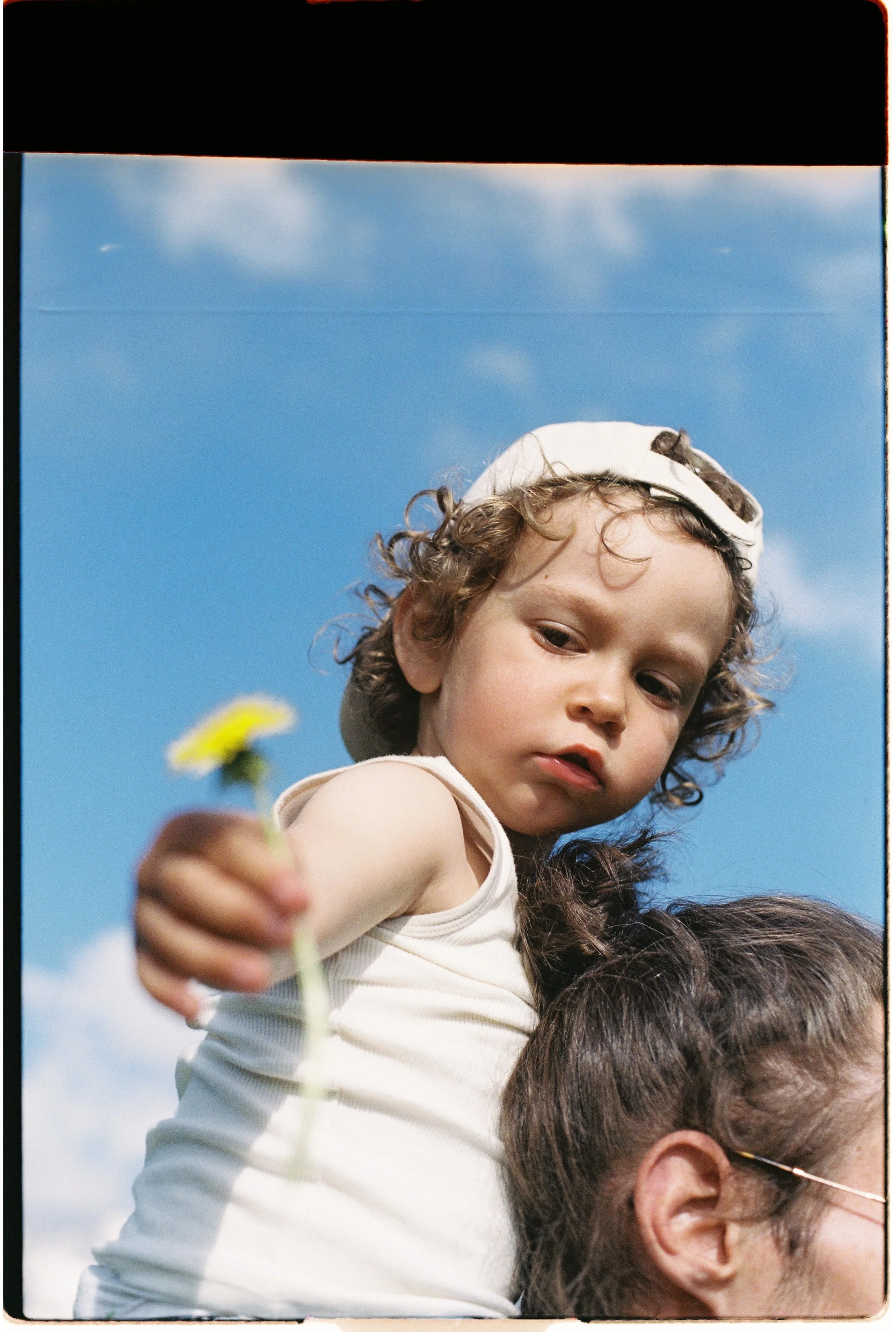 Une jeune fille avec des cheveux bouclés porte un chapeau blanc, elle regarde vers l'objectif et tient une fleur blanche dans sa main, tandis qu'une partie d'une personne adulte est visible en dessous d'elle, sous un ciel bleu avec quelques nuages.