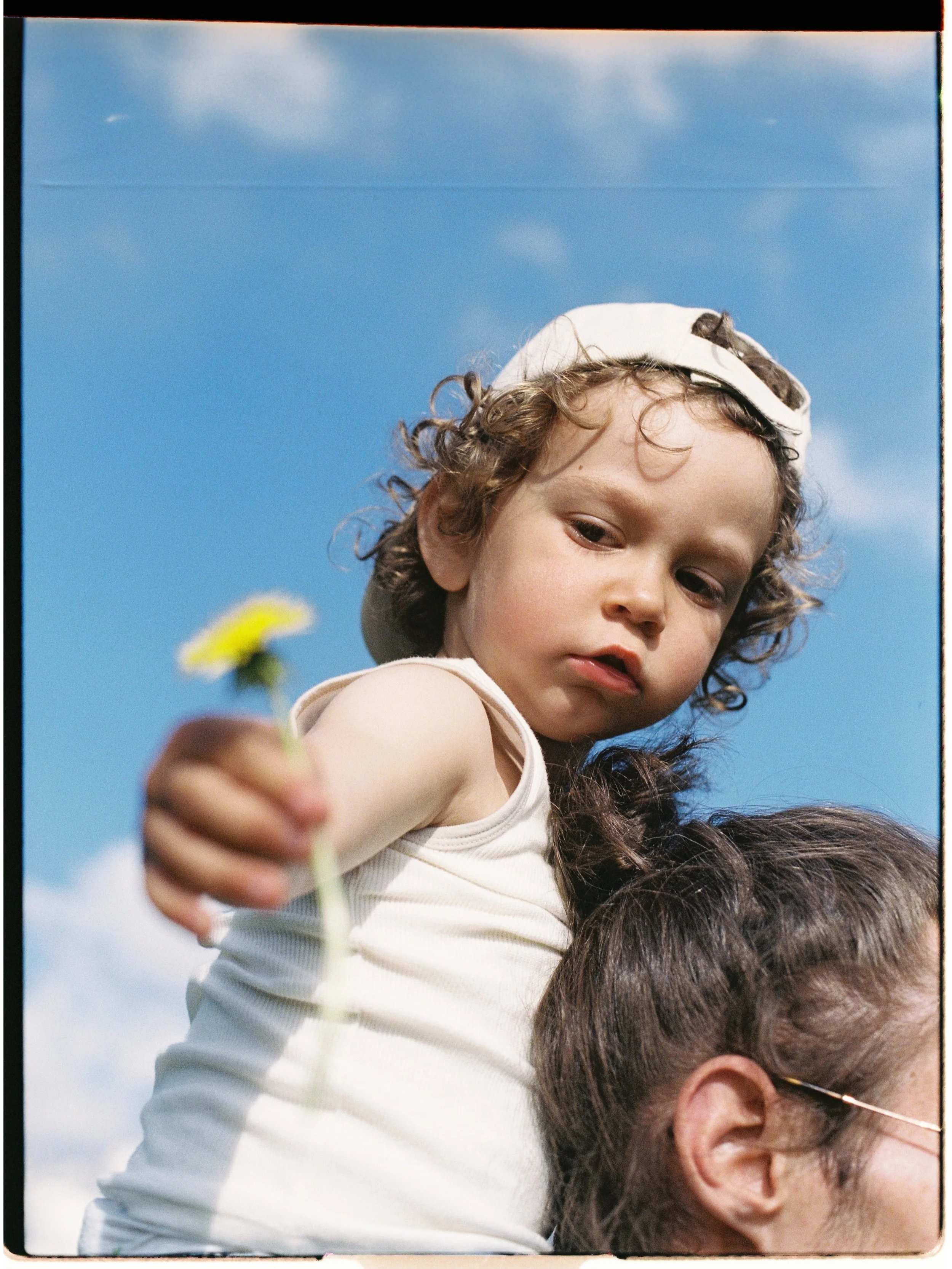 Un enfant blond avec une casquette blanche est assis sur les épaules d'une personne dont seule la tête avec des cheveux bruns et des lunettes est visible, contre un ciel bleu avec quelques nuages.