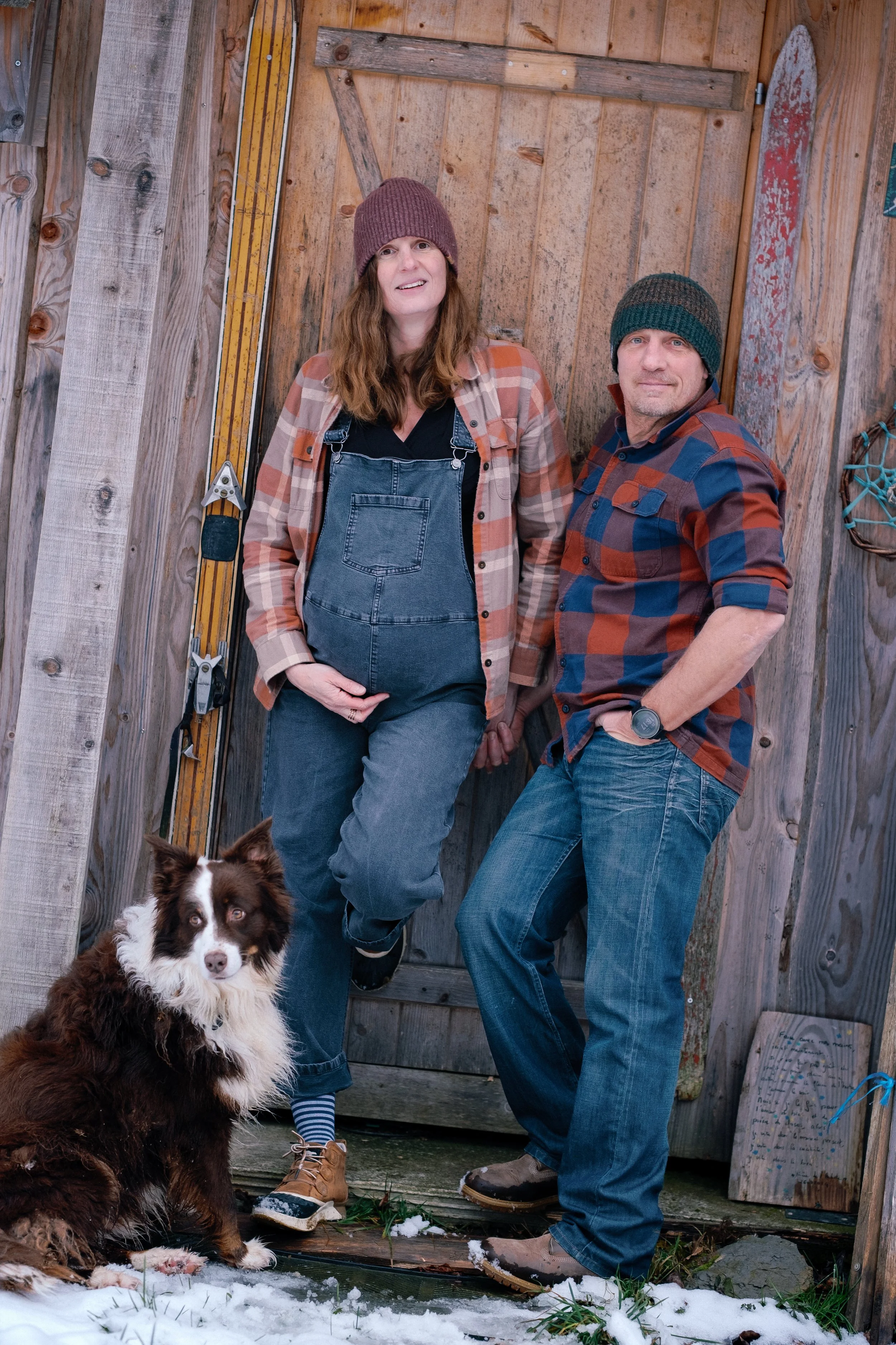 Un couple pose devant une porte en bois avec leur chien de race Border Collie dans un environnement extérieur enneigé.