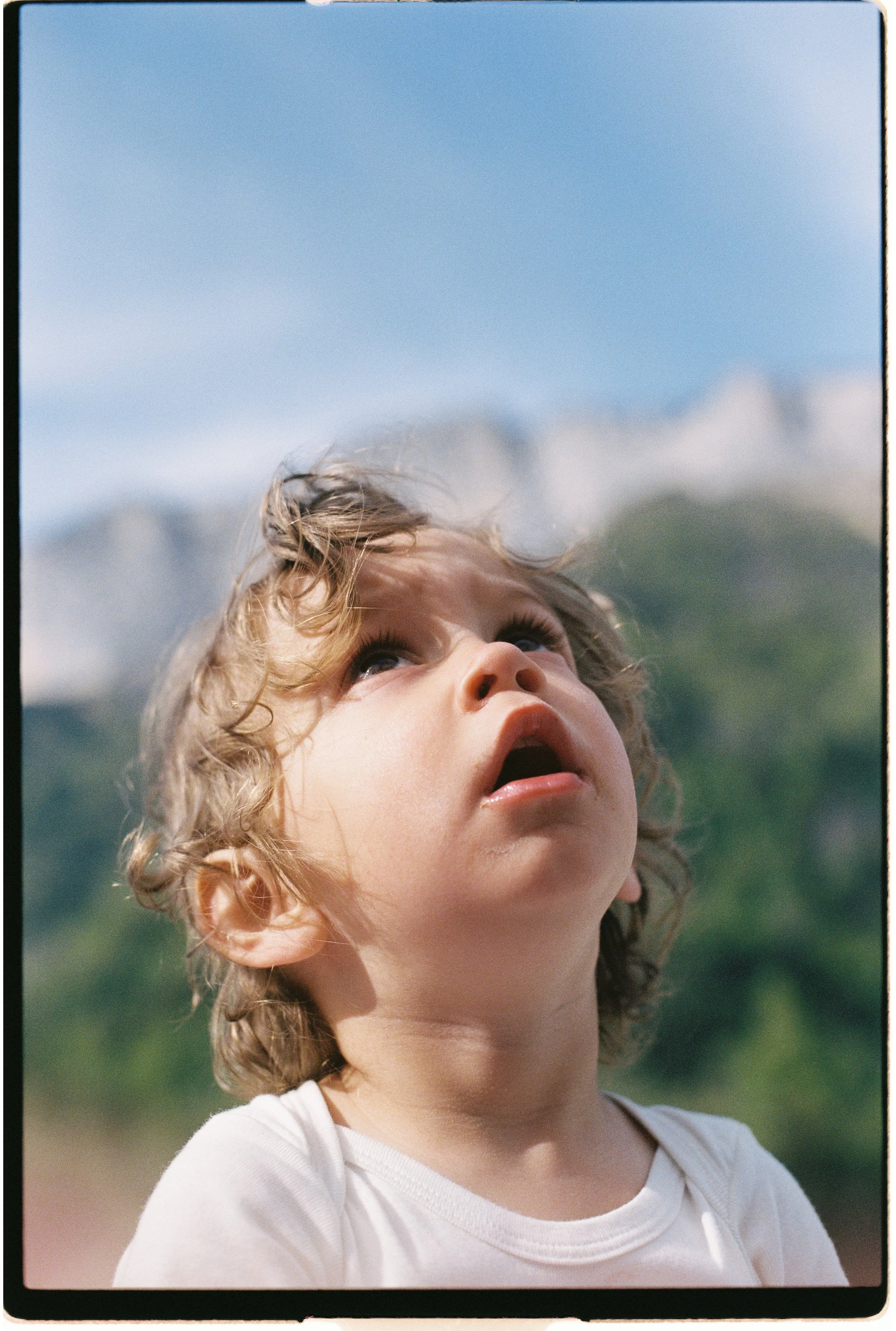 Jeune fille aux cheveux bouclés, regards vers le haut, portant un t-shirt blanc, en plein air, ciel bleu en arrière-plan.