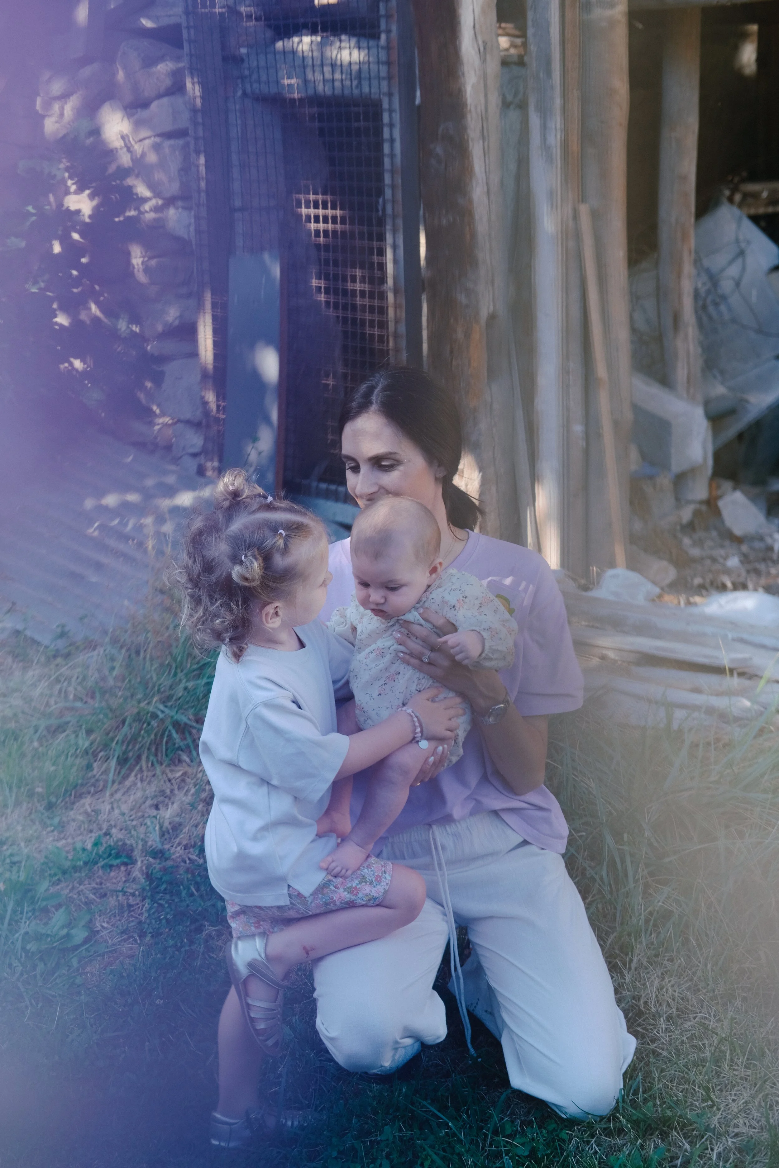 Une femme mère tenant deux enfants, un garçon et une bébé, en plein air avec des structures en bois et des matériaux de construction en arrière-plan.