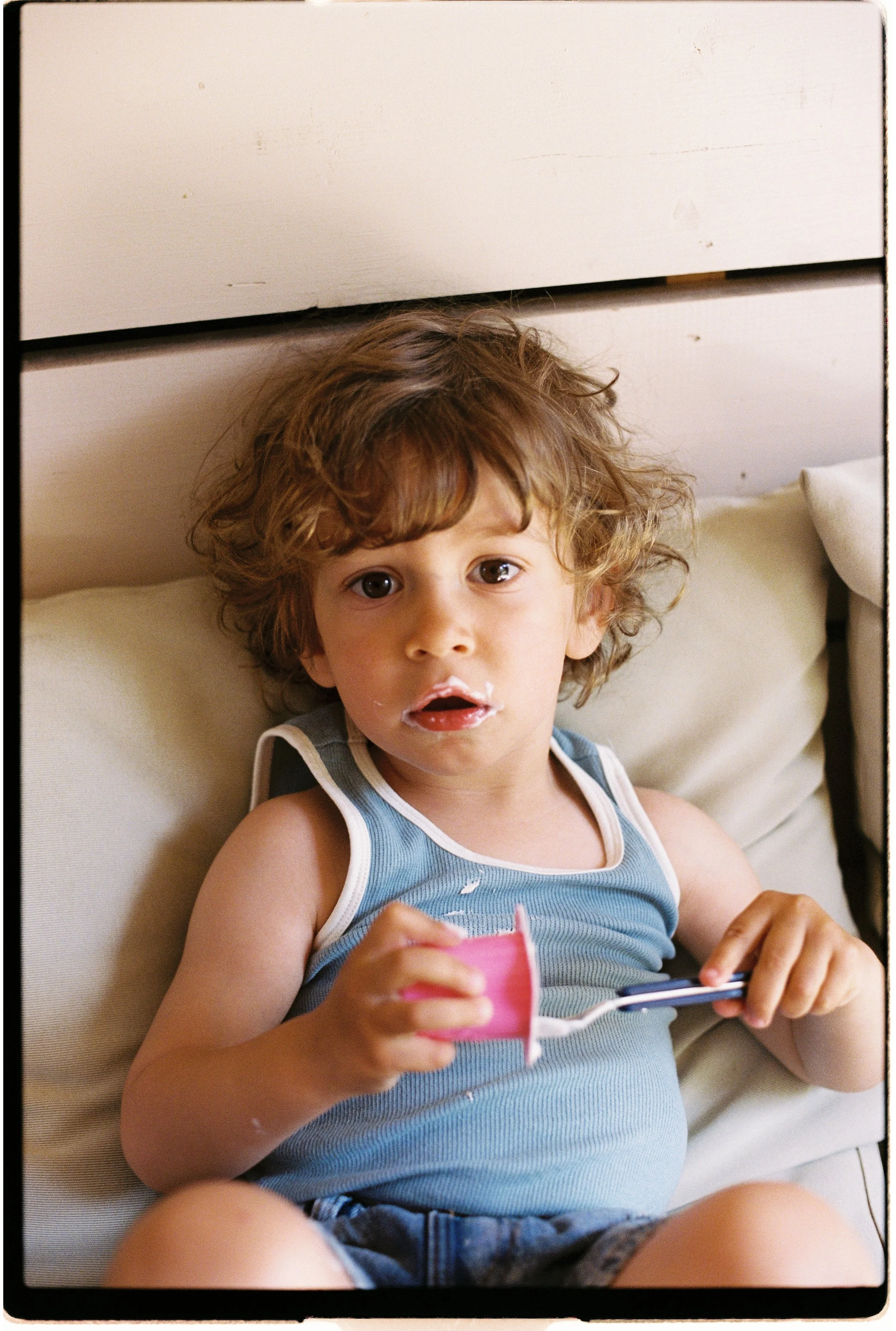 Un jeune garçon aux cheveux bouclés et aux yeux marron clair, allongé sur un lit avec un coussin beige, tenant un petit suisse. Il en a autour de la bouche et semble surpris ou curieux.