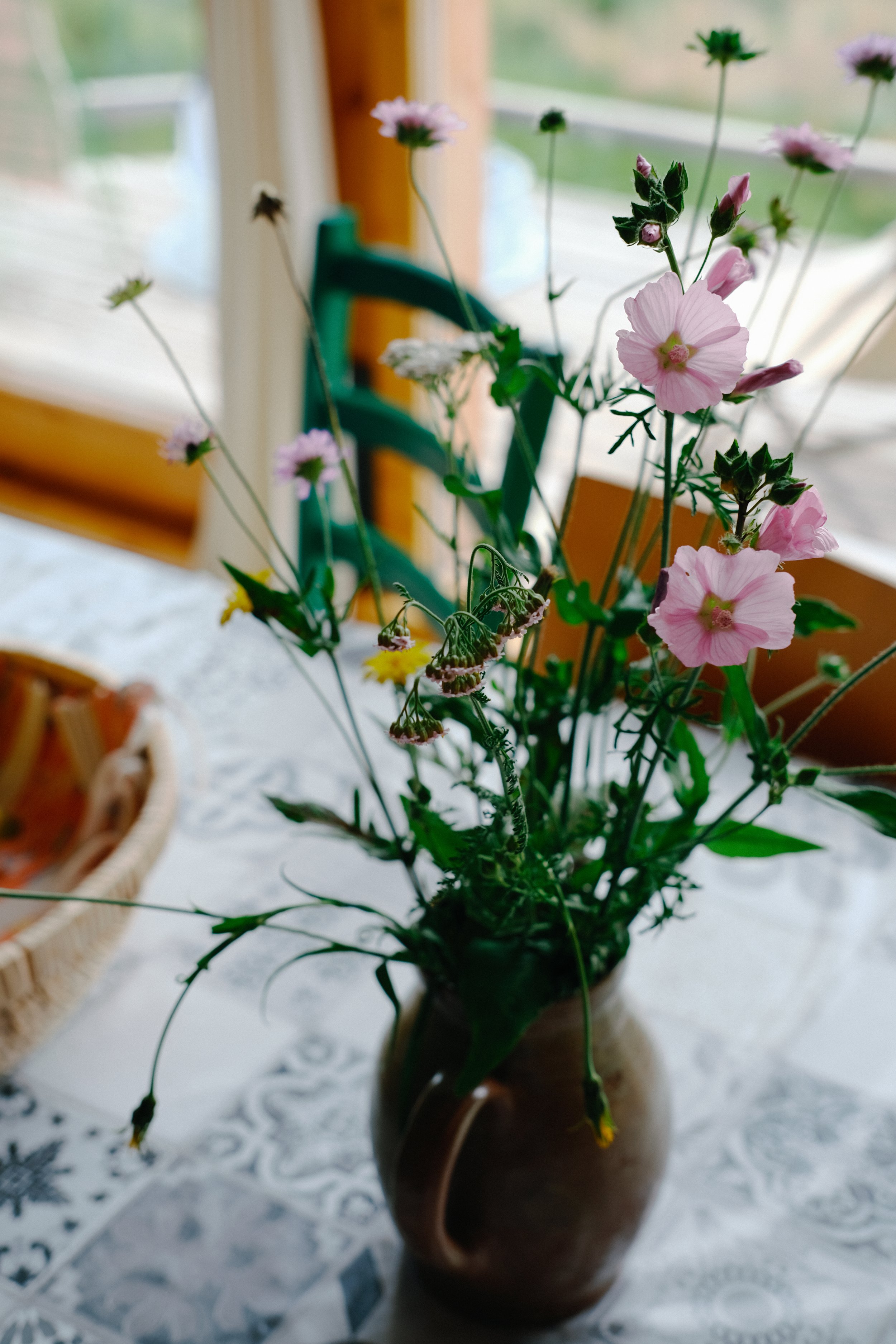 Un bouquet de fleurs dans un vase en terre cuite placé sur une table avec une nappe blanche, avec une fenêtre en arrière-plan.