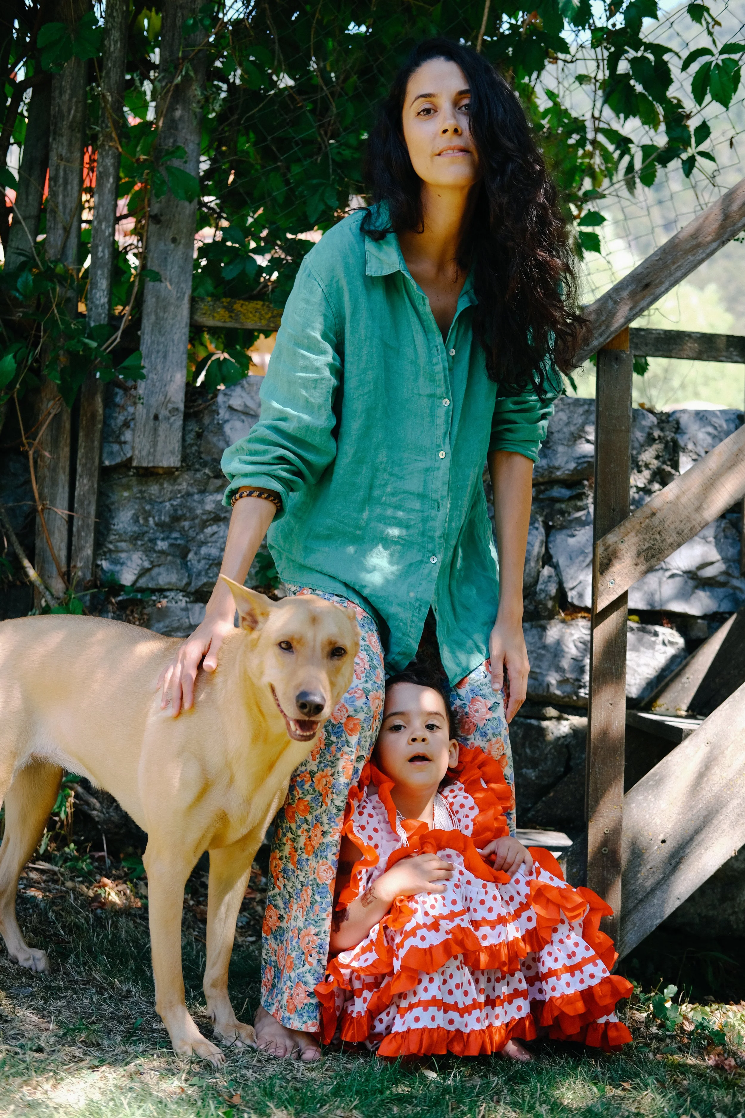 Une femme avec des cheveux bouclés noirs portant une chemise turquoise, deux enfants, un garçon en robe à pois rouge et blanc, et une fille en surplis orange et blanc, ainsi qu'un chien beige, posent dans un environnement naturel avec des arbres et u
