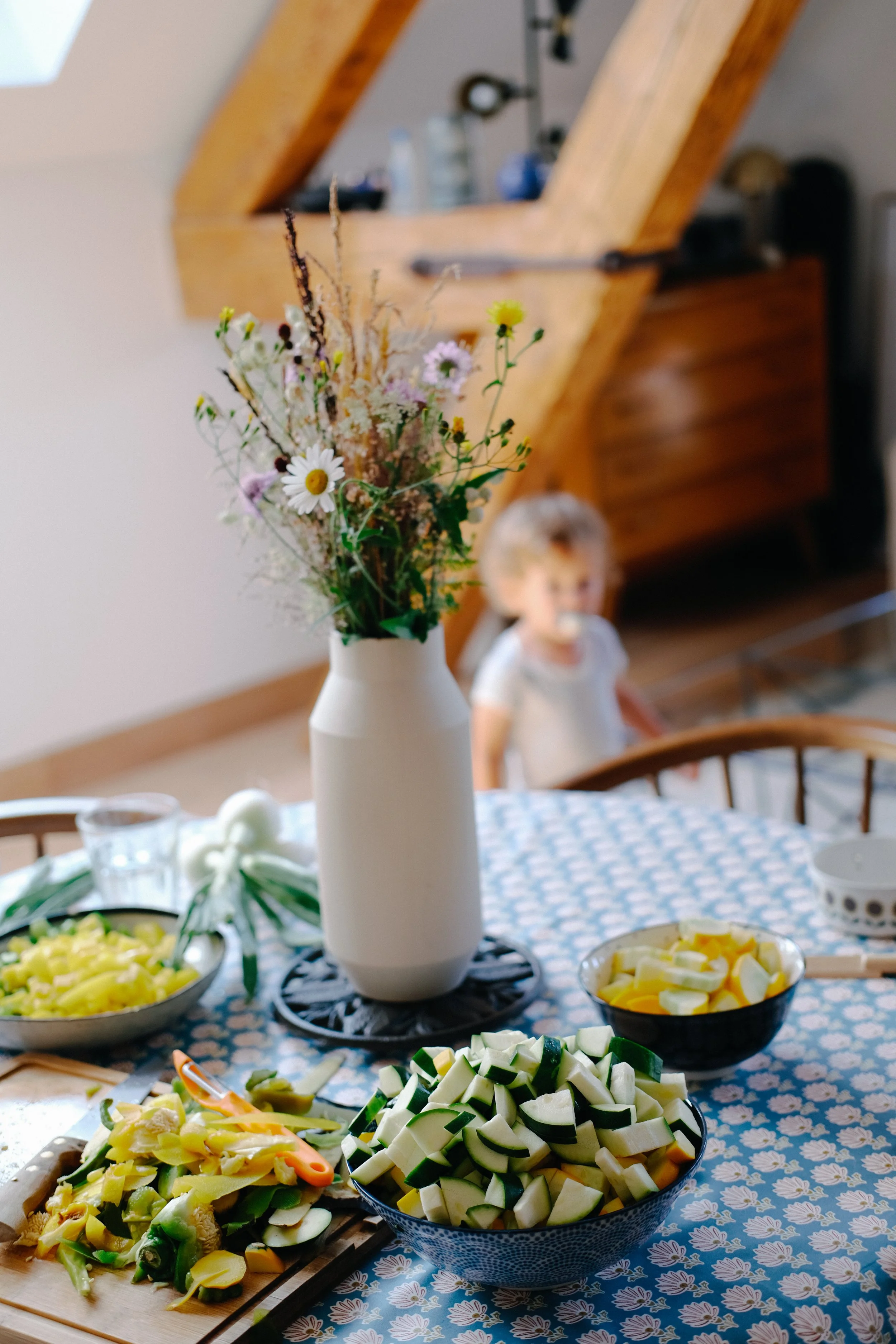 Vase blanc avec un bouquet de fleurs sauvages, table avec des salades et légumes coupés, décor floral sur la nappe, ambiance conviviale et chaleureuse.