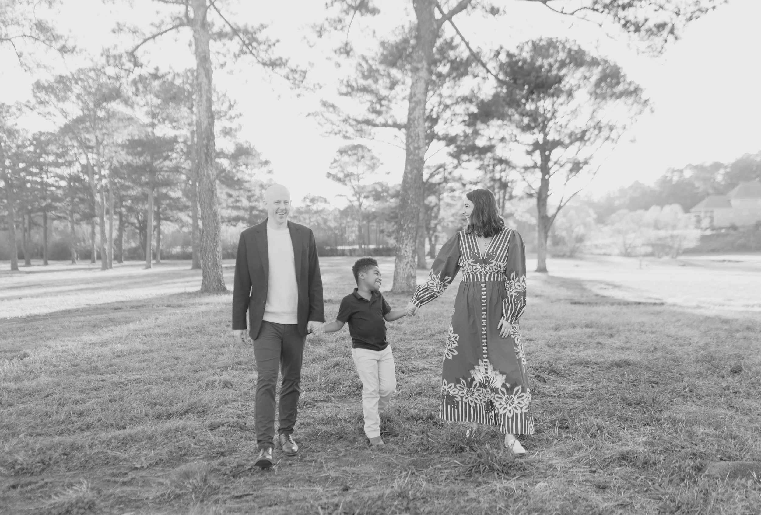 A family of three, a man, woman, and young boy, walking hand-in-hand through a park with trees in the background. The family is smiling and enjoying each other's company.