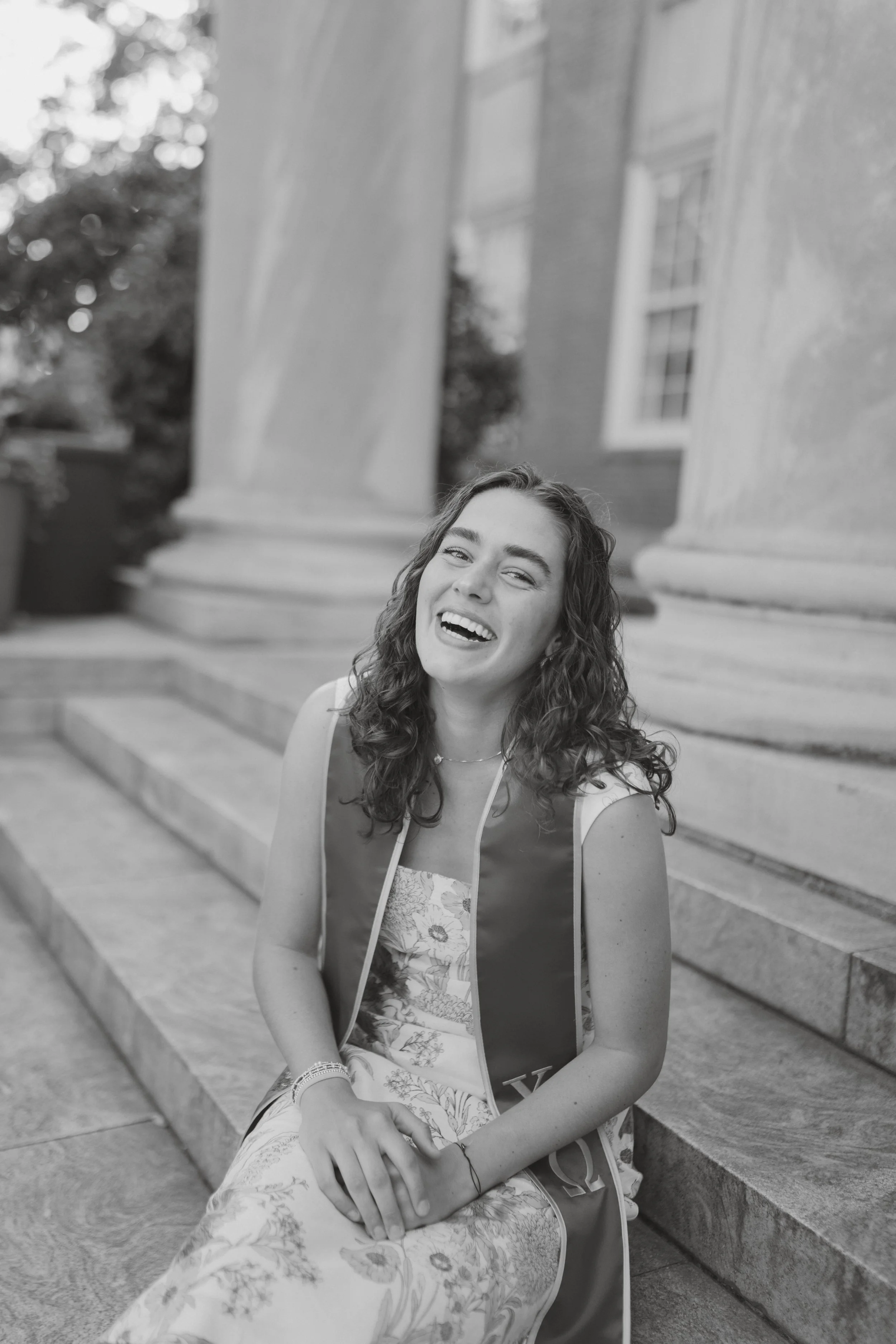 Black and white photo of a young woman with curly hair, sitting on stone steps in front of a building with columns, smiling and wearing a graduation stole over a floral dress.