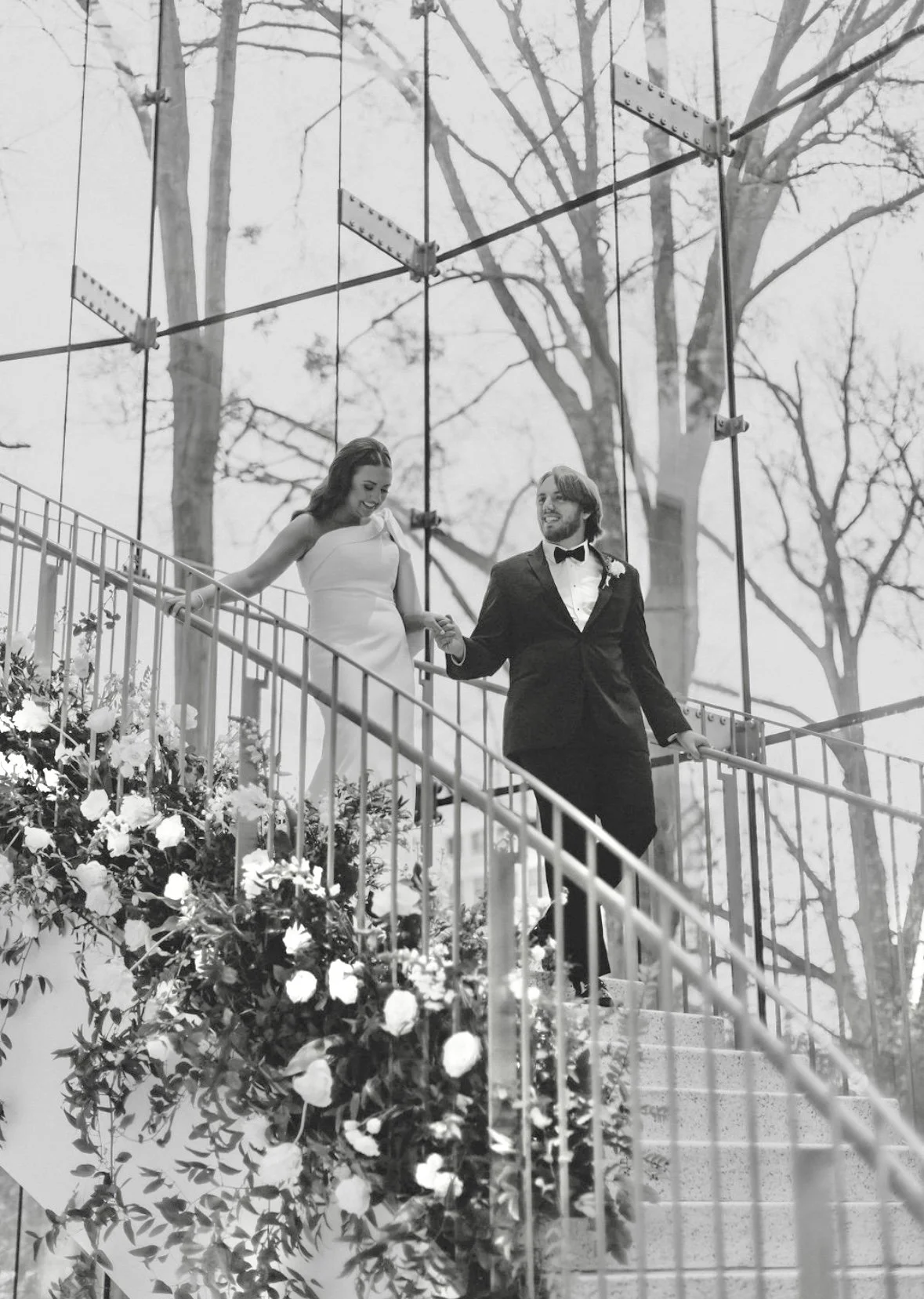 A bride in a white dress and a groom in a black tuxedo walking down a staircase decorated with flowers, holding hands and smiling.