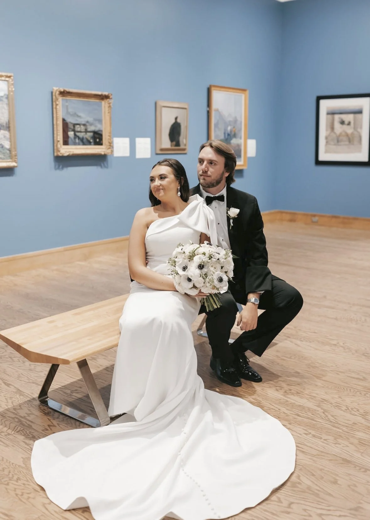 A bride and groom sitting on a wooden bench inside an art gallery, with framed paintings on the blue wall behind them.