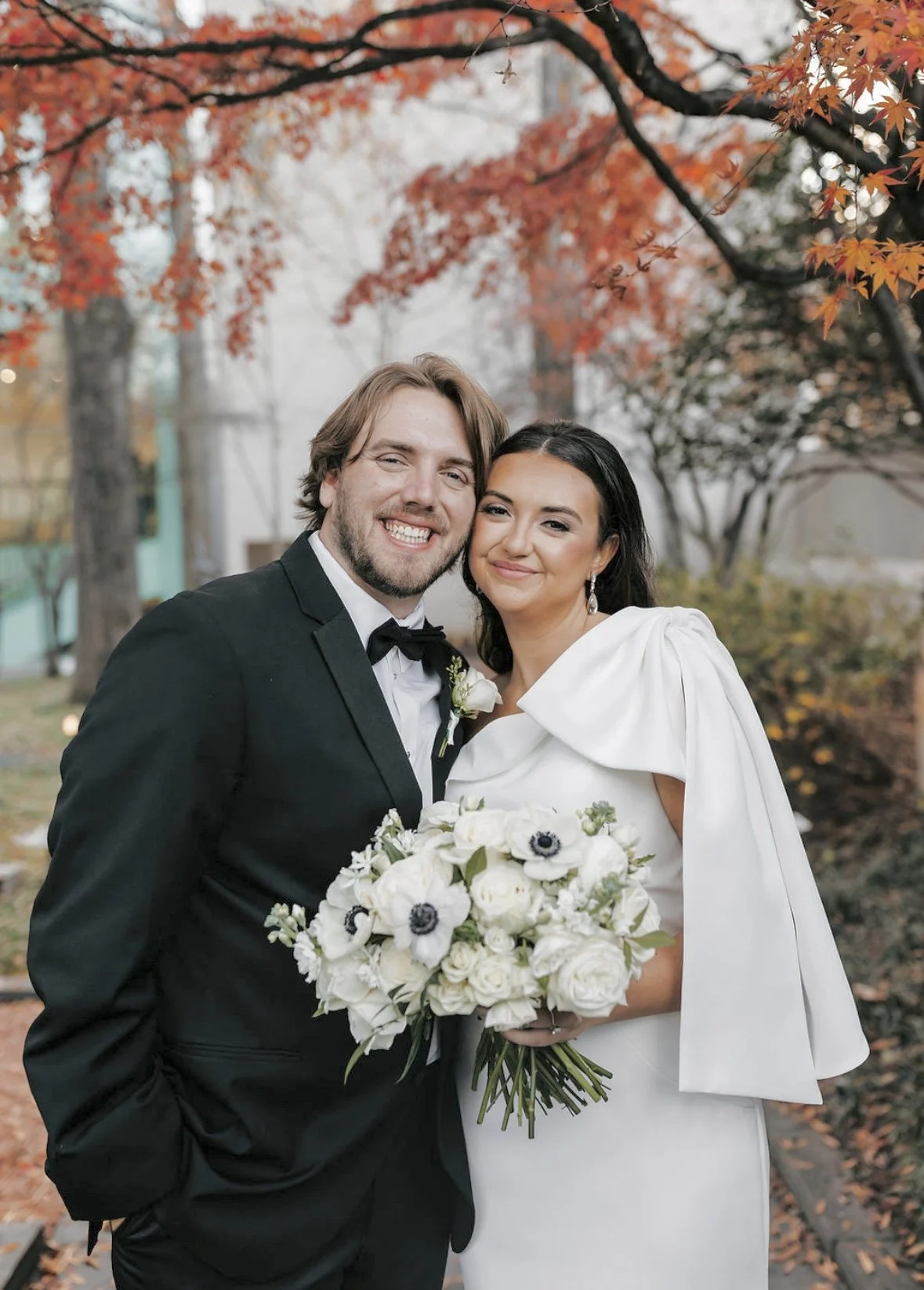 A newlywed couple smiling and standing close together outdoors in a park with autumn orange and red leaves, the groom in a black tuxedo with a bowtie and a white boutonniere, and the bride in a white dress holding a bouquet of white flowers and anemo