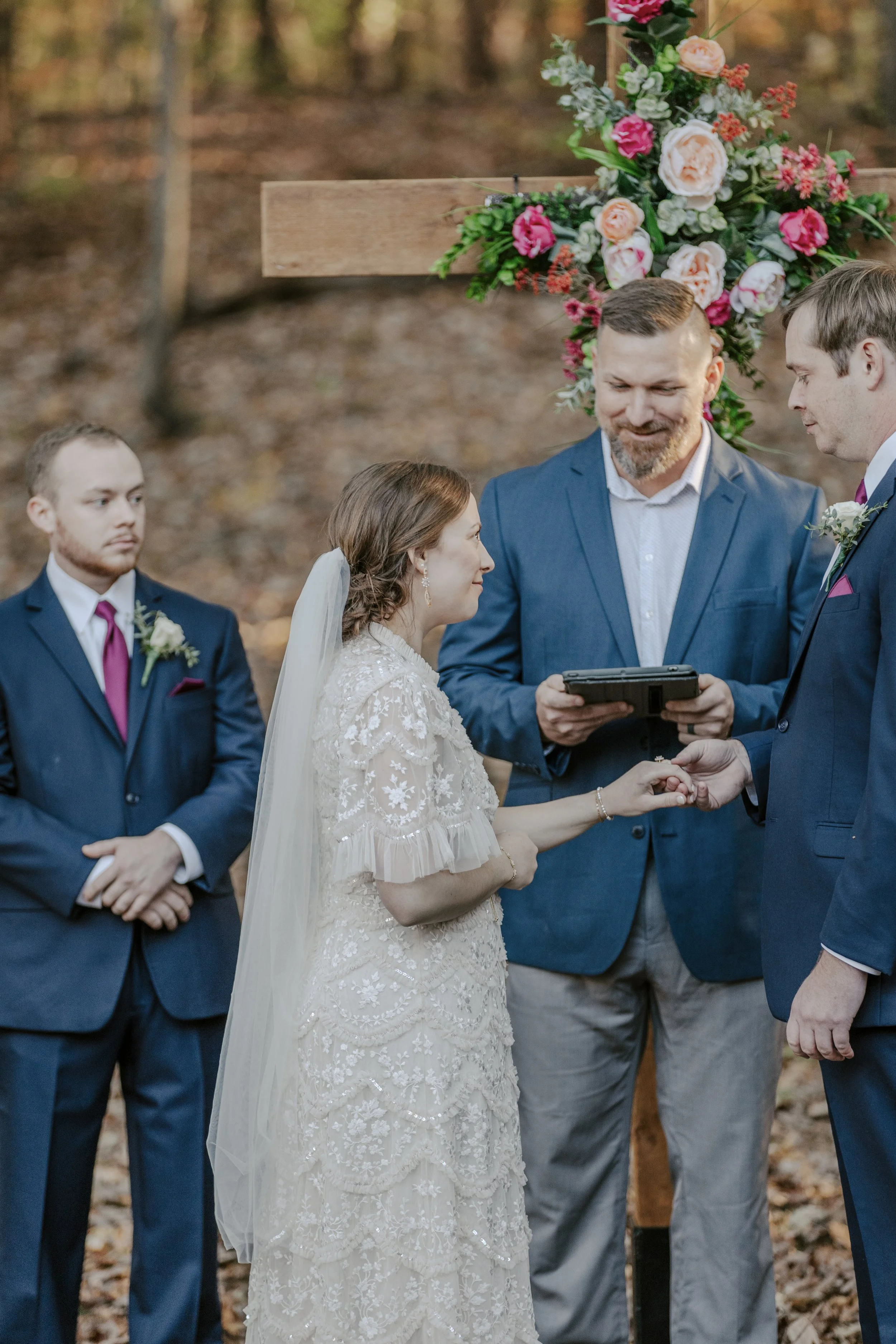 A couple getting married outdoors, holding hands, with an officiant and two groomsmen, surrounded by trees and decorated with a floral arrangement behind them.