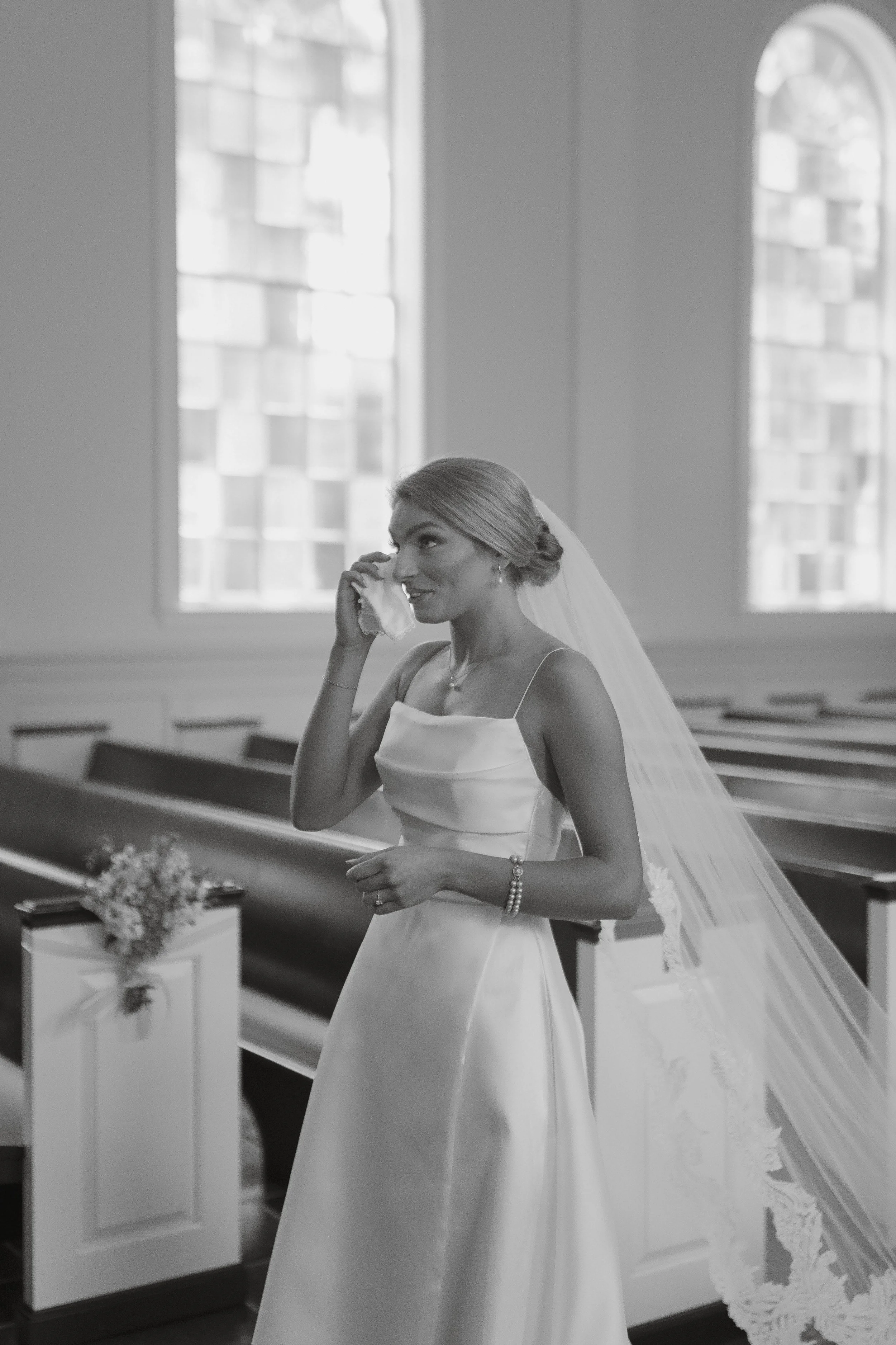 A bride in a satin wedding dress with thin straps, standing inside a church, adjusting her veil while smiling.