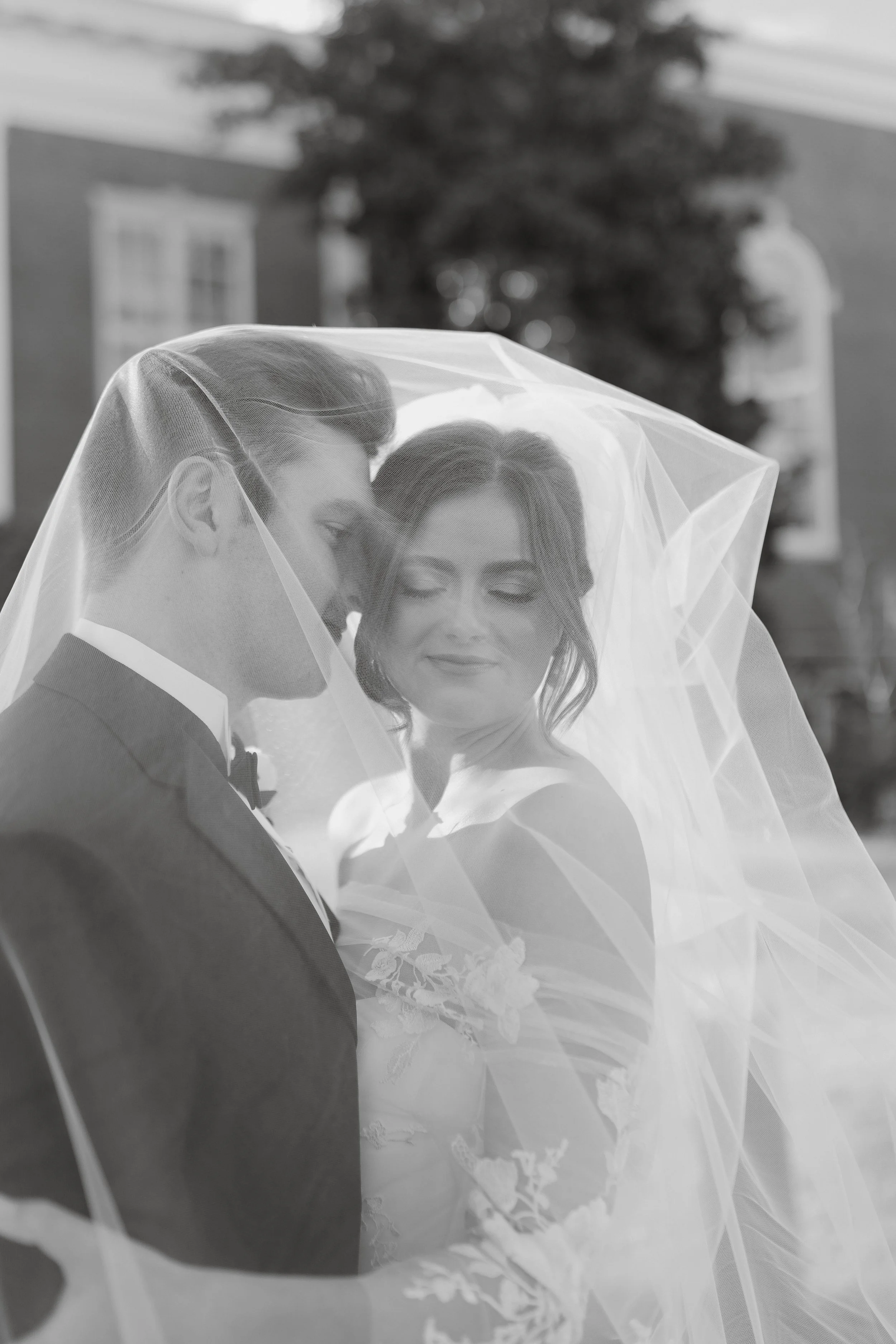 Black and white photo of a bride and groom under a veil, with the groom wearing a suit and bow tie, and the bride in a wedding dress holding a bouquet, standing outdoors with blurred buildings in the background.