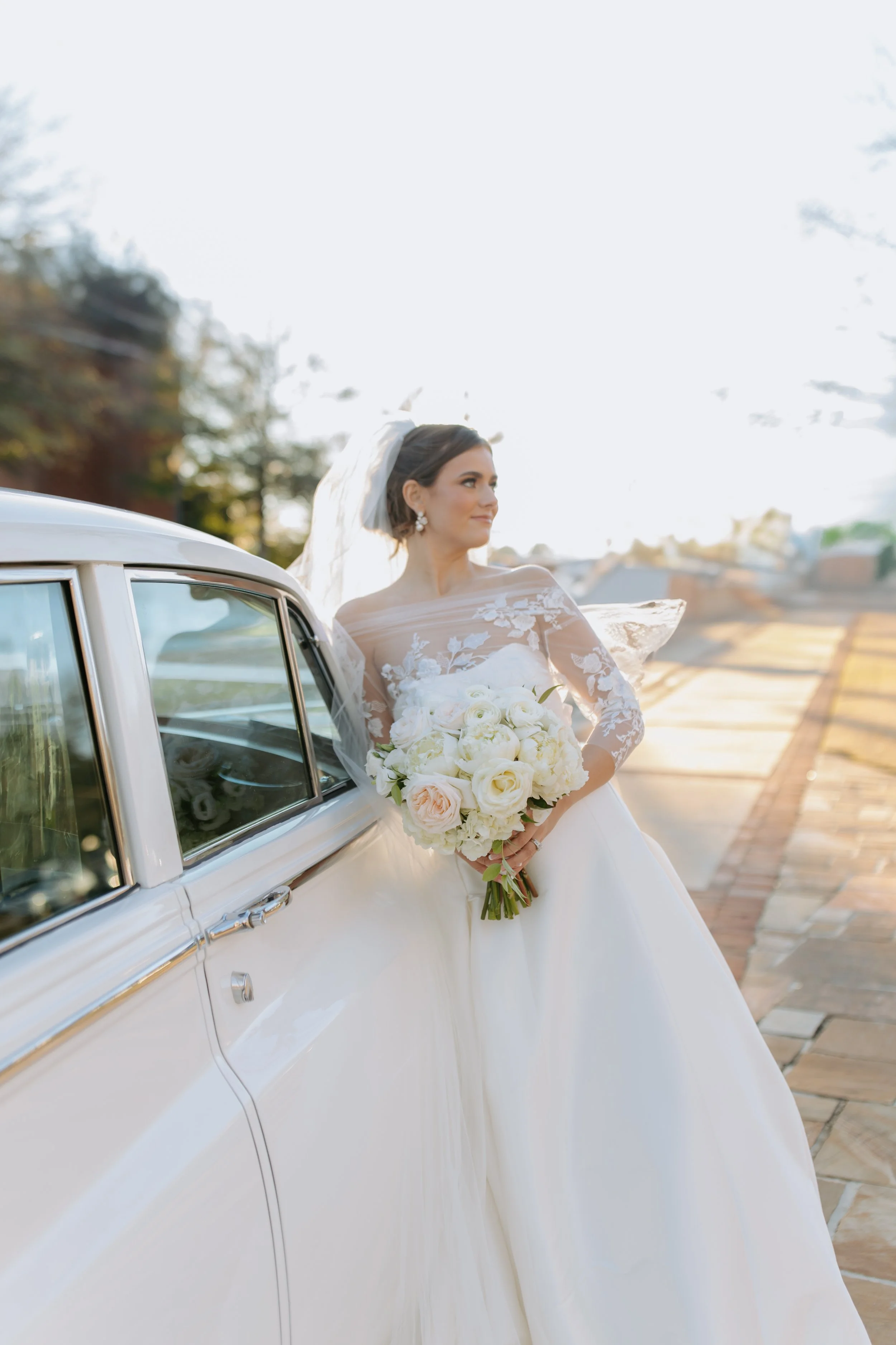 Bride in a white wedding dress holding a bouquet of white and blush roses, leaning against a vintage white car outdoors during sunset.