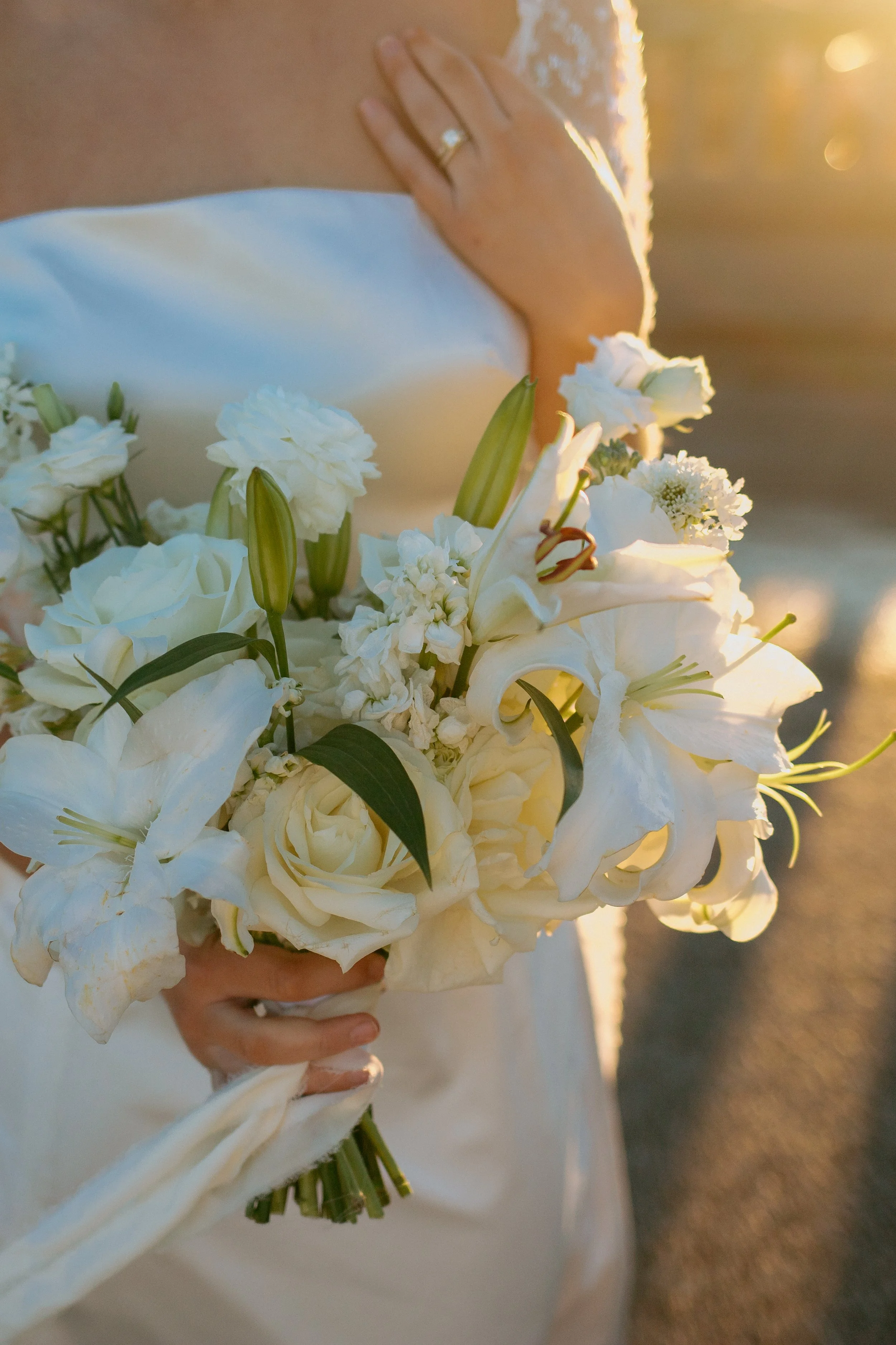 A bride holding a bouquet of white flowers including lilies and roses, with her wedding ring visible on her left hand during sunset.