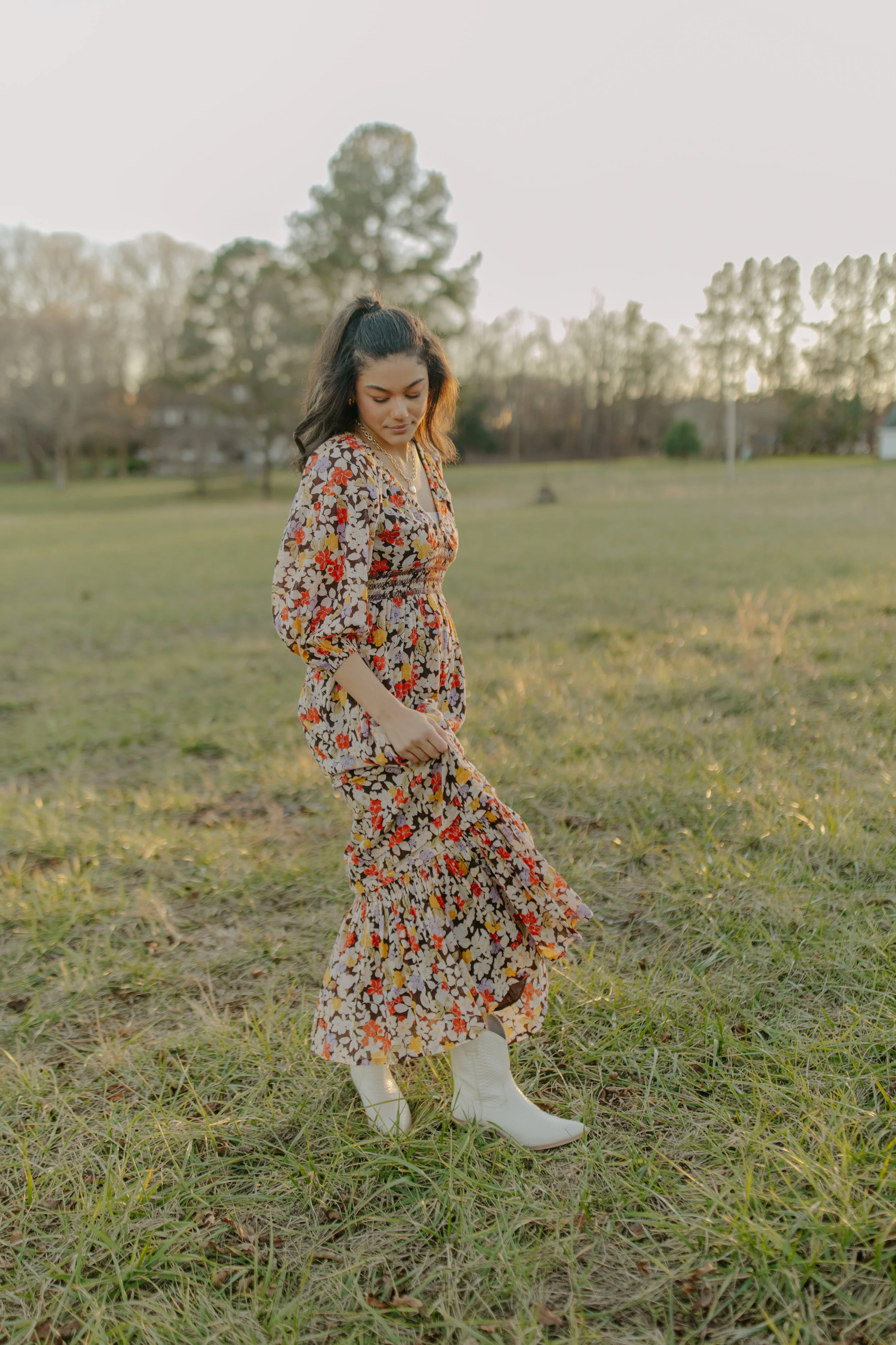 Woman wearing a floral dress and white cowboy boots standing in a grassy field during sunset.