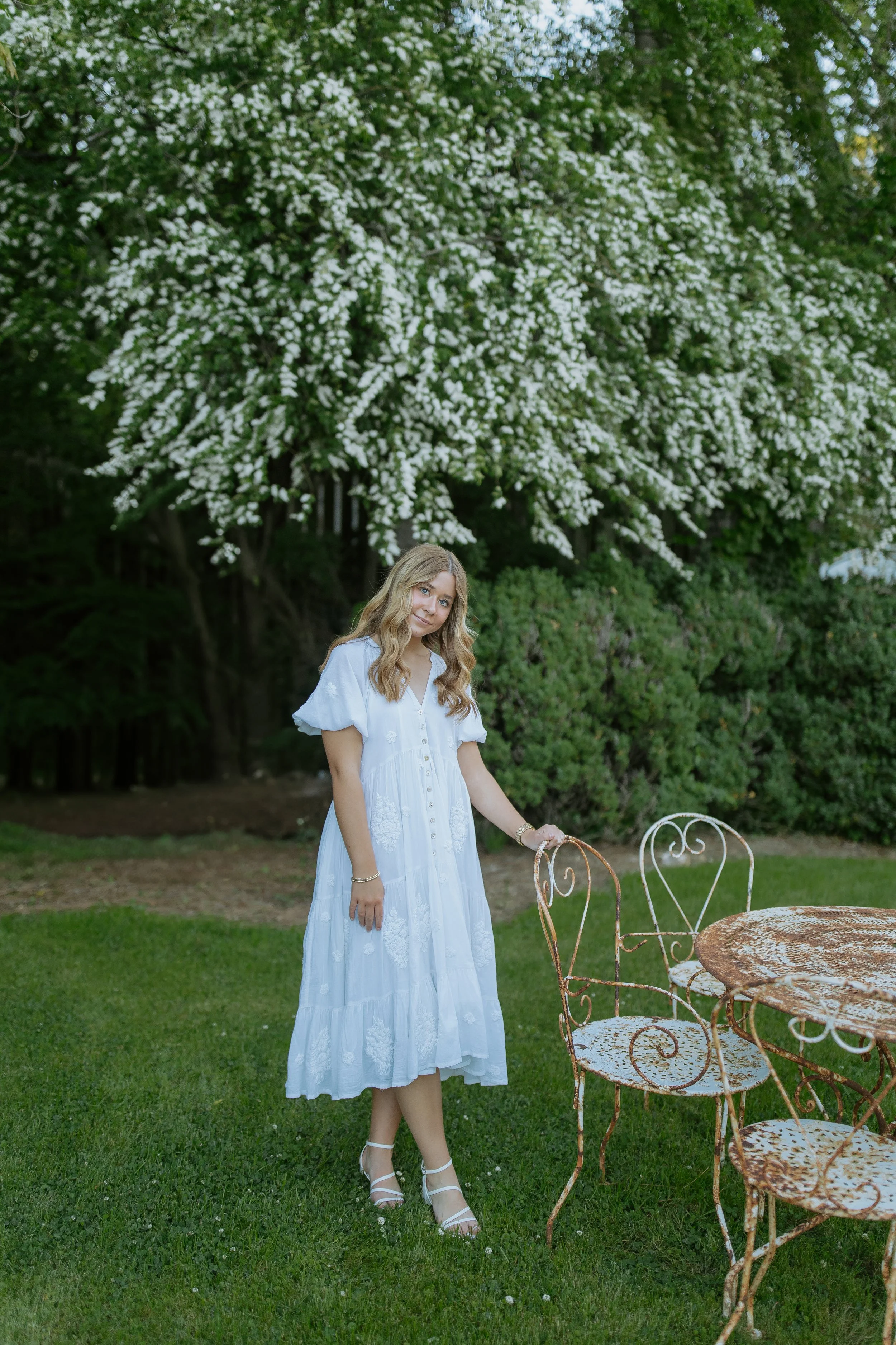 Young woman in a white dress with embroidered details standing next to a rusty, antique iron patio table and chairs on a green lawn in a garden with a large blossoming tree in the background.