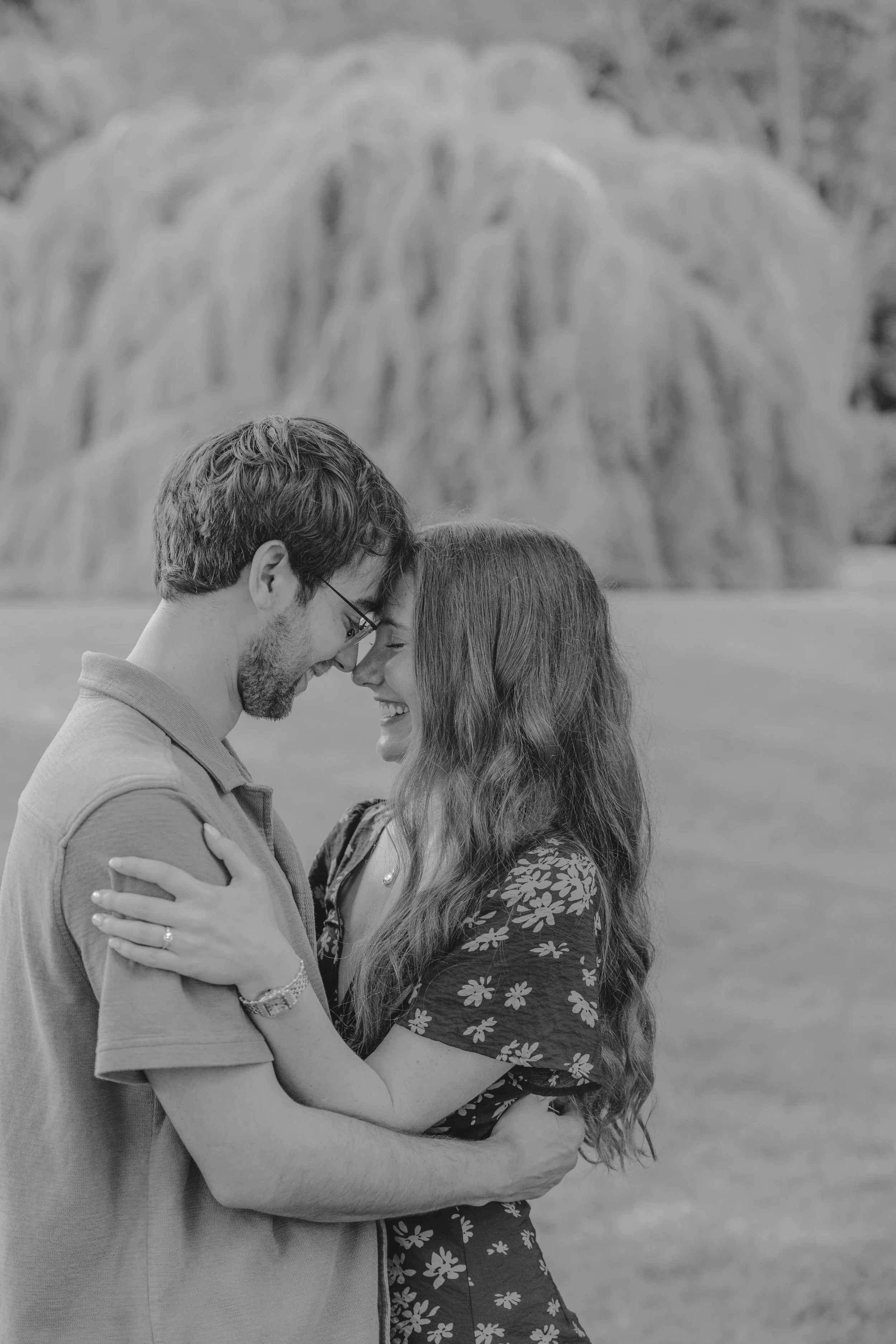 A black and white photo of a couple smiling and embracing each other with their foreheads touching, outdoors with a large rock formation in the background.