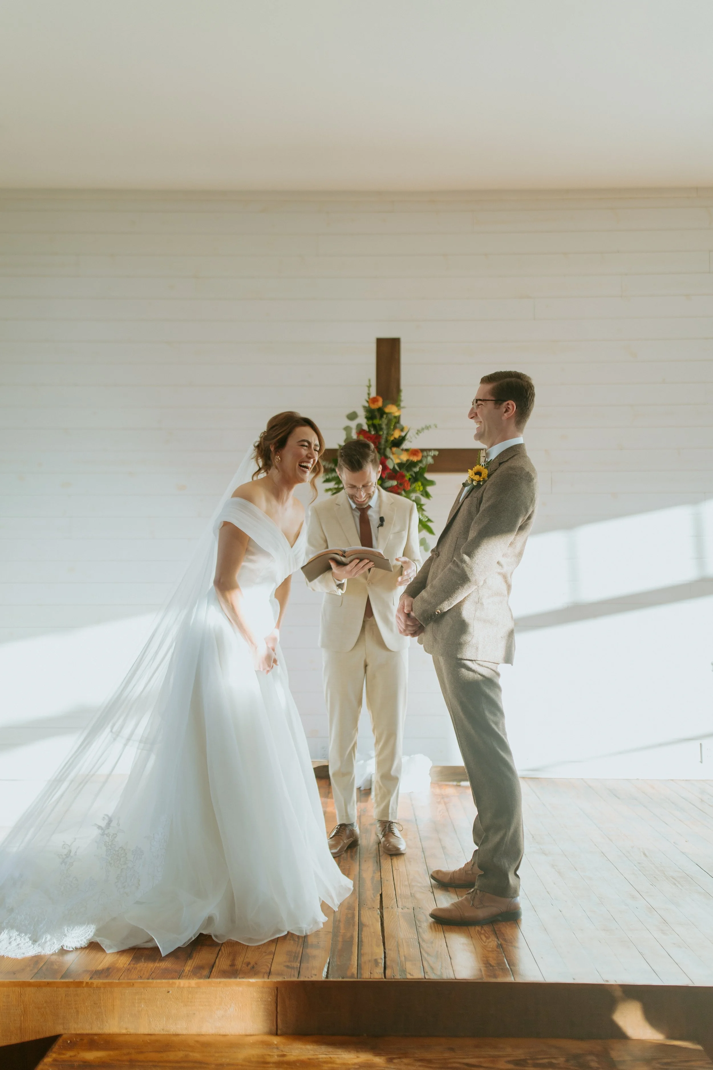 A bride and groom standing at the altar during their wedding ceremony, with a officiant reading from a book. They are both smiling and laughing. There is a flower arrangement and a cross in the background, with sunlight streaming in.
