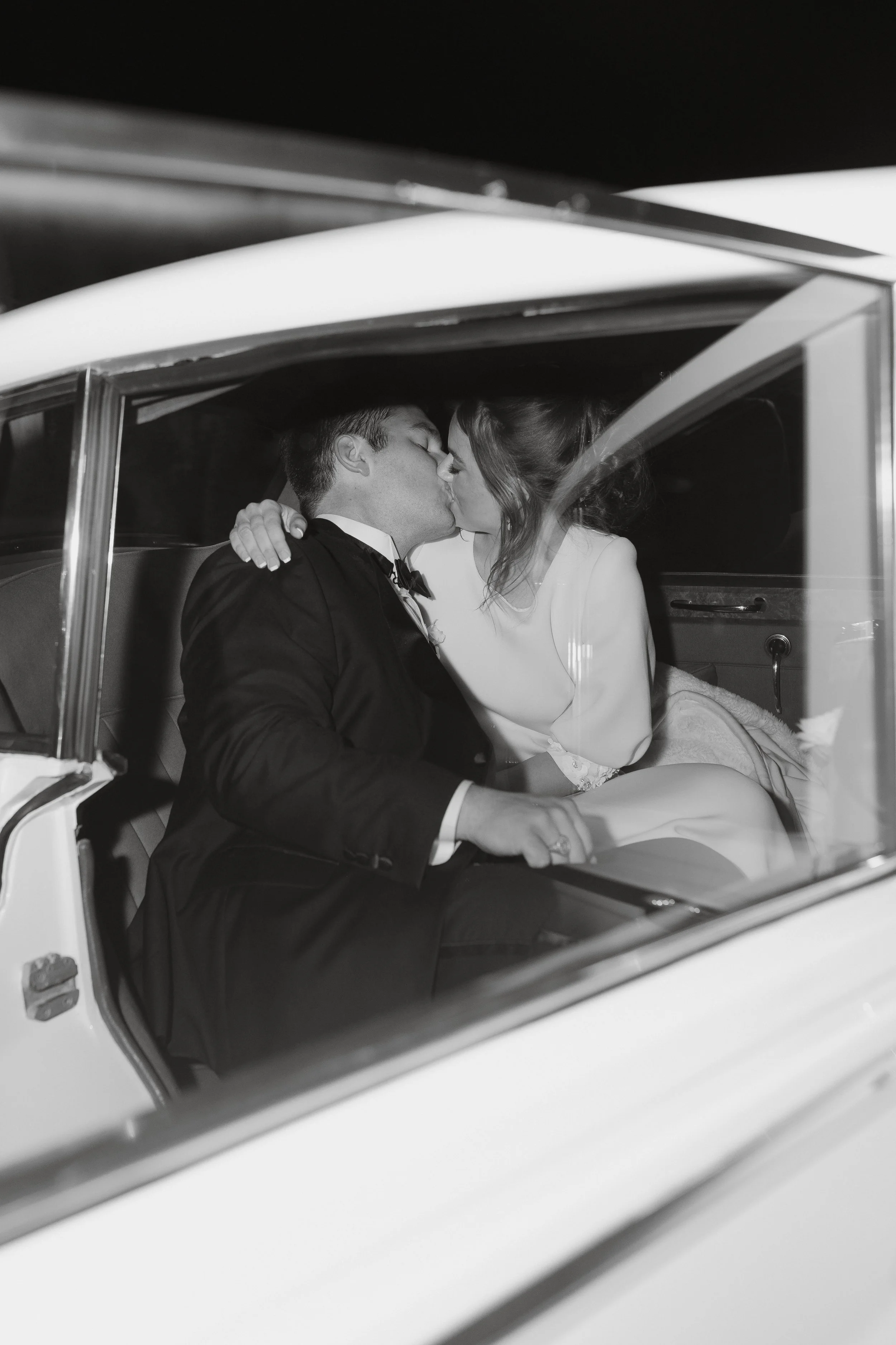 Black and white photo of a newly married couple sharing a kiss while sitting in the backseat of a vintage car, romantic and intimate moment captured at night.