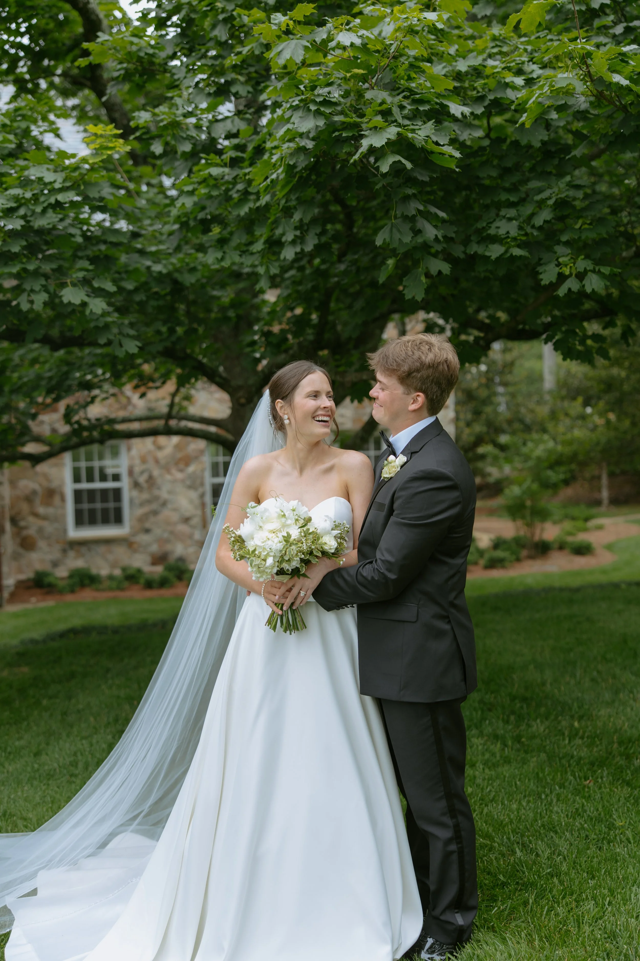 A bride and groom on their wedding day, standing outdoors under a large leafy tree, smiling at each other, with a stone building in the background.